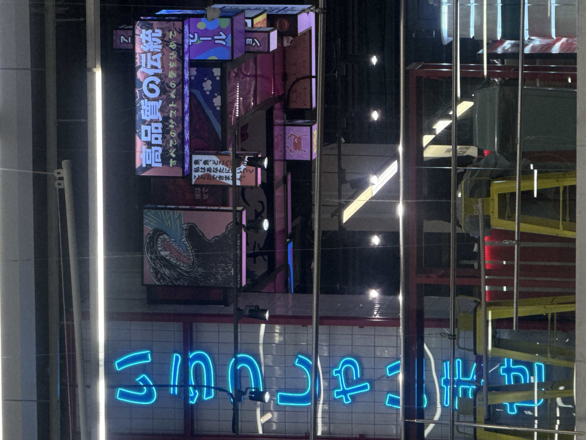 The image depicts an indoor commercial establishment with a prominent Japanese aesthetic, located in Vladivostok, Russia. The scene is dominated by artificial lighting, including vibrant neon signs and illuminated display panels, against a generally dark background.
On the left side of the frame, a vertical blue neon sign is displayed against a white tiled wall. The sign spells out 「いらっしゃいませ」 (Irasshaimase) in hiragana, which translates to "Welcome." Below this, a portion of the sign continues, reading 「ませ」 (mase).
Towards the center and right, a complex arrangement of illuminated signs and panels creates a collage effect. A large horizontal sign at the top displays 「高品質の伝統」 (Kōhinshitsu no dentō), meaning "Tradition of High Quality," with a smaller subtitle below it that reads 「すべてのゲストへの愛を込めて」 (Subete no gesuto e no ai o komete), meaning "With love for all guests." To the left of this sign, a poster features a black and white illustration of Godzilla roaring against a pink background.
Below these larger displays, a multi-tiered structure comprises several smaller illuminated boxes. One visible box on the far right displays 「ビール」 (Bīru), meaning "Beer." Another panel shows a pink crab silhouette on a dark background. Other panels feature abstract patterns or partially visible Japanese text, including phrases like 「魚、魚、どこへいく」 (Sakana, sakana, doko e iku), meaning "Fish, fish, where are you going," and parts of sentences implying questions or statements about actions.
In the midground, metal railings or banisters separate the viewer's perspective from the main area. Behind these railings, several yellow bar stools are visible, positioned in front of a counter area that features red accent lighting. A large, dark gray bin or container is also present in the midground. Overhead, fluorescent lights and smaller track lights illuminate specific areas. No discernible individuals are clearly visible or engaged in specific actions within the frame. The overall impression is that of a modern, themed restaurant or food court setting.