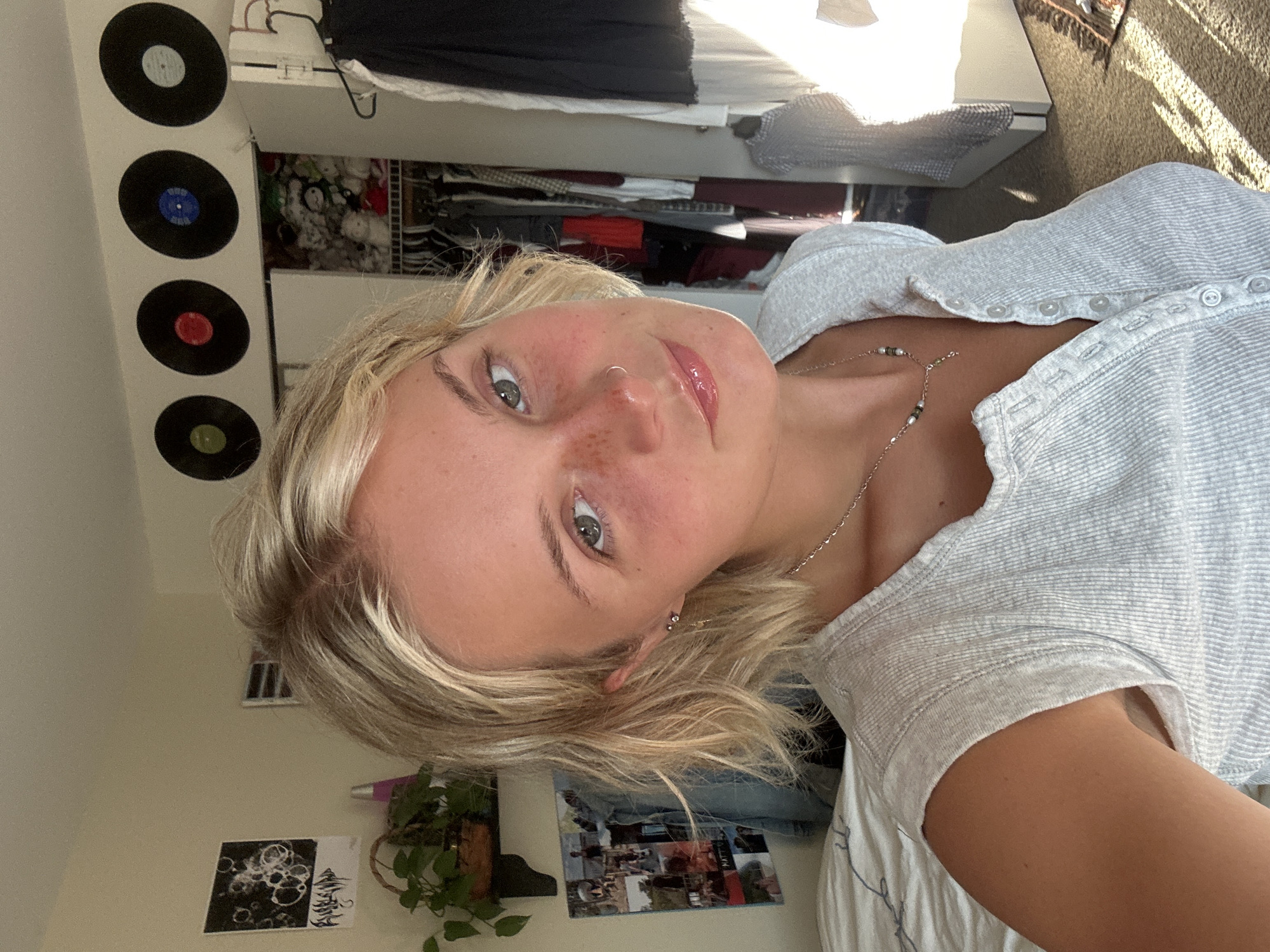 The image shows a young woman with blonde, wavy hair and light skin with freckles. She is wearing a light grey, ribbed, button-up shirt and a delicate silver necklace with small beads. She has a nose ring and a small earring. Her makeup appears natural with lip gloss. She is looking towards the camera with a slight smile.
In the background, the setting appears to be a bedroom. There are several vinyl records displayed on the wall above a closet filled with clothing and stuffed animals. A shelf with a plant is visible to the left, along with a collage of photos. A bed with white, patterned bedding is in the foreground on the left. Sunlight is streaming in from the right, casting long shadows on the floor and illuminating the scene. The overall impression is a casual, personal space. The location is not identifiable as Whistler, Canada.