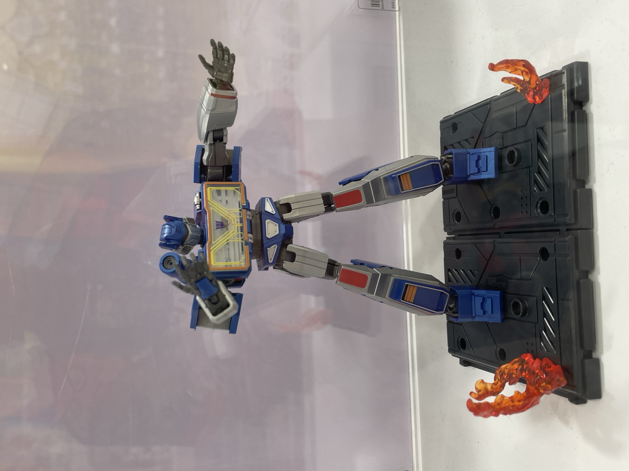 A full-body shot of a Soundwave transformer action figure stands on a black display base. The figure is predominantly blue and silver with red accents on its legs and chest. Its left arm is bent at the elbow, with a gun-like accessory pointed forward. Its right arm is extended outwards. The display base has two red flame-like accessories on either side. The background is a blurred, light purple-toned surface, suggesting it is behind a glass enclosure. There are no people visible in the image. The scene appears to be indoors, possibly in a retail or exhibition setting. There are no discernible landmarks or features that would indicate a specific location within Davao City, Philippines.