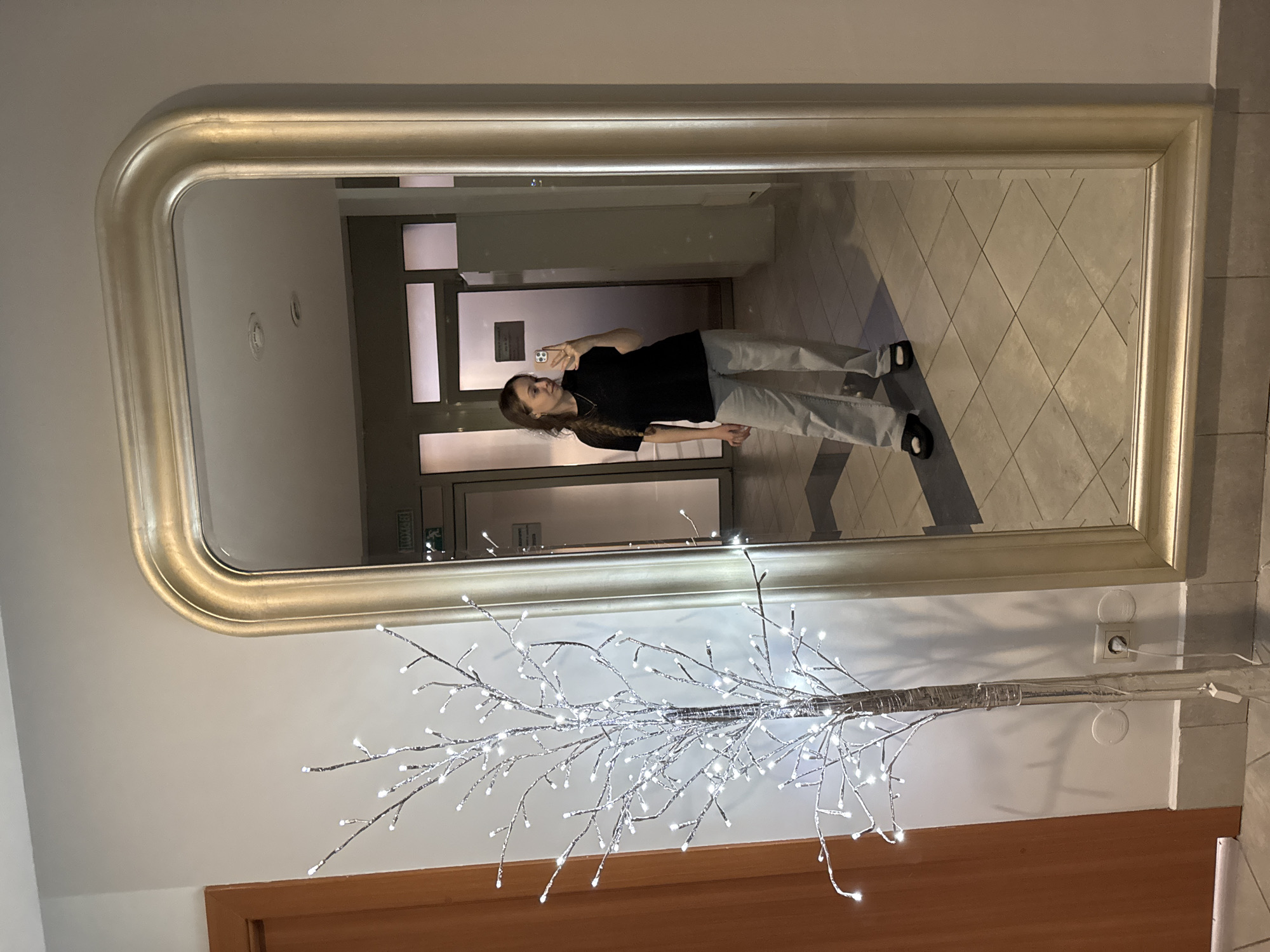 A young woman is taking a mirror selfie indoors. She has long brown hair and is wearing a black shirt and light-colored pants. The mirror has a rounded silver frame. A decorative light-up tree is situated below the mirror. The mirror reflects a hallway with tile floors and closed doors. A sign with Cyrillic text and an exit symbol is visible above one of the doors in the reflection. The flooring in the reflection has a tile pattern. An electrical outlet is located near the decorative tree. The location is likely a building in Yekaterinburg, Russia.