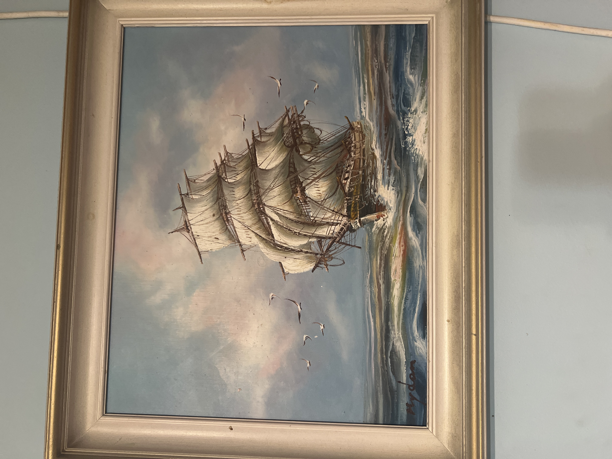 A framed oil painting depicts a tall ship sailing on a choppy sea under a cloudy sky. The ship, with multiple sails set, is shown from the port side, appearing to move towards the viewer. Several seagulls fly in the sky around the ship. The sea is painted with dynamic brushstrokes, showing whitecaps and waves. The sky is a blend of blue, pink, and white, suggesting a dramatic atmosphere. In the bottom left corner of the painting, a signature is visible in dark red paint, which reads "Hyden". The painting is housed in a light-colored, textured frame with a thin gold inner border. The background wall appears to be painted a light blue.