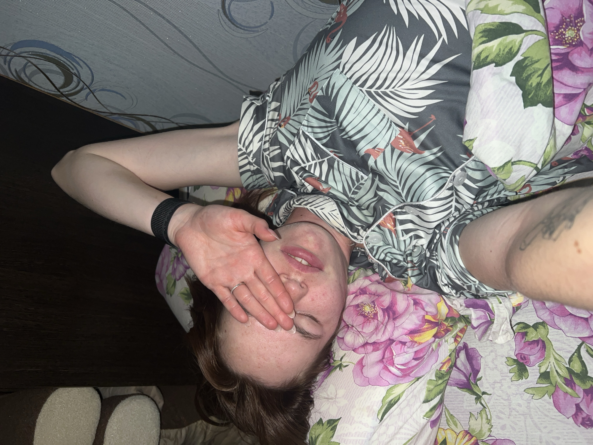The image shows a young Caucasian woman lying on a bed, covering her eyes with her right hand. She is wearing pajamas with a tropical pattern featuring palm leaves and flamingos. The bed is covered with floral-patterned bedding. The woman has reddish-brown hair and appears to be asleep or drowsy. The background includes a dark headboard and a wall with a textured wallpaper pattern. The overall lighting suggests it is nighttime or a dimly lit room. There is no visible indication of the location within the city of Chaikovsky, Russia.