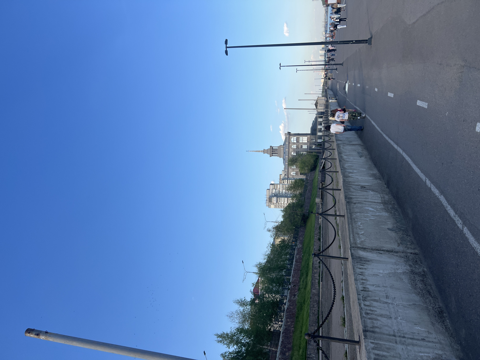 The image shows a daytime view of a section of a street or embankment in Krasnoyarsk, Russia. The sky is clear and blue. The road is made of dark asphalt. A white painted line marks a lane on the road. People are present on the right side of the image, walking along the embankment. There are tall lamp posts along the road. On the other side of the road is a concrete wall with a metal chain fence. Trees and grass grow behind the wall. Buildings, including one with a distinctive spire, are visible in the background.