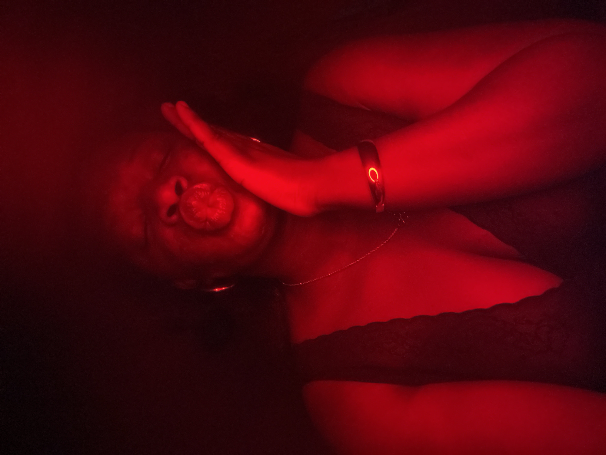 A person is lying down with their face partially obscured by their hand. The lighting is predominantly red, creating a dramatic effect. The person is wearing what appears to be a black lace top. They also have jewelry, including a bracelet and a necklace. The environment is dark and indistinct. The image location is not detectable.