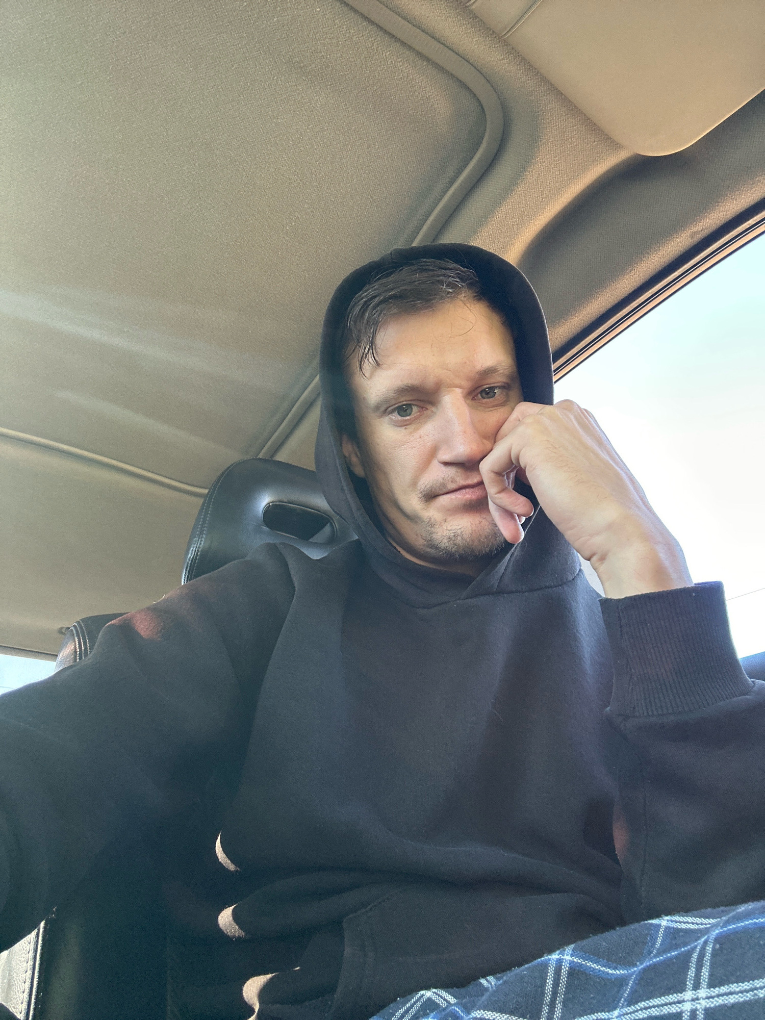 A man wearing a black hooded sweatshirt and plaid pants is seated inside a vehicle. His right hand is resting on his chin, and he is looking downwards with a neutral expression. The interior of the car appears to be beige, with sunlight streaming in from the windows. The scene is a close-up shot, focusing on the man's face and upper body. There is no discernible location in the city of Makiv, Ukraine.