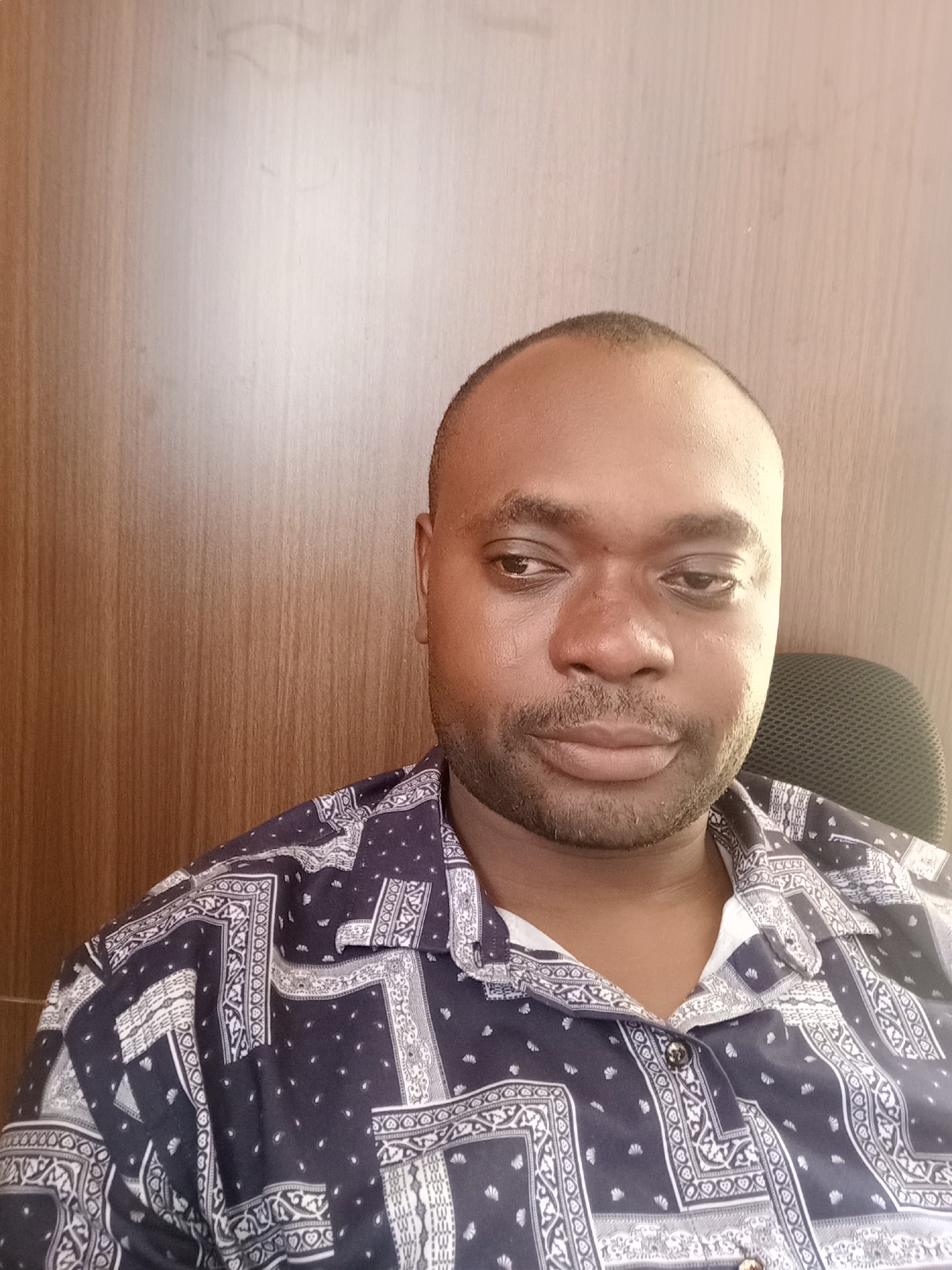 A close-up shot shows a man seated in what appears to be an office setting. He has dark skin, a shaved head, and a neatly trimmed beard. He is wearing a button-down shirt with a dark blue and white paisley-like pattern. Behind him is a wood-paneled wall and the back of a black mesh office chair is partially visible to his right. The setting is indoors, suggesting an office or home office environment. There is no visual evidence to confirm the location is in Gwarinpa, Nigeria.