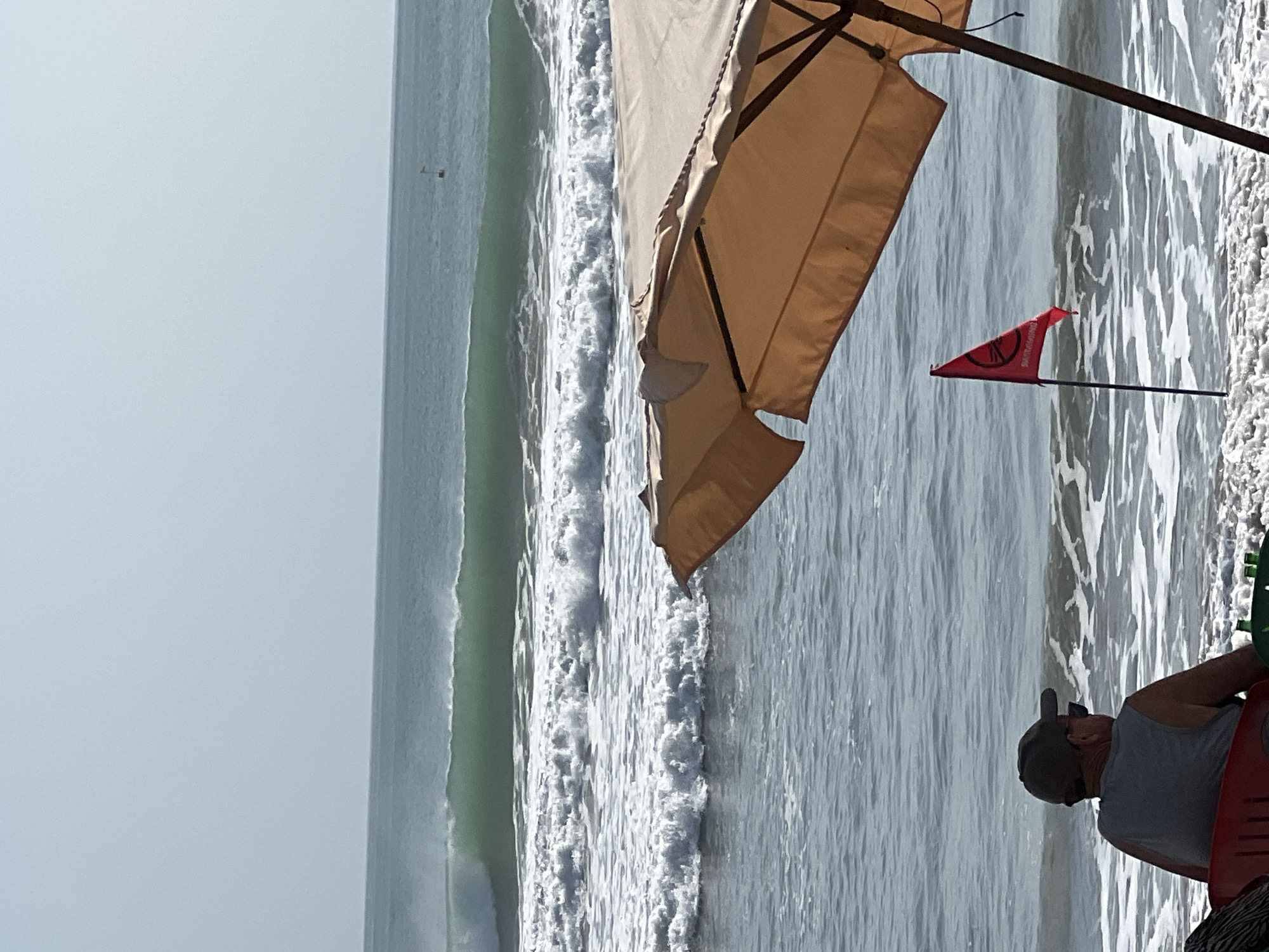The image depicts a serene beach scene in Kuta, Indonesia. A person wearing a gray tank top and a baseball cap is seated on a red chair, facing the ocean. In the foreground, a tan beach umbrella shades part of the beach, and a red flag with white lettering, possibly indicating swimming conditions, is planted in the sand. The ocean exhibits gentle waves, with a larger wave forming in the distance. The sky is clear and pale blue.