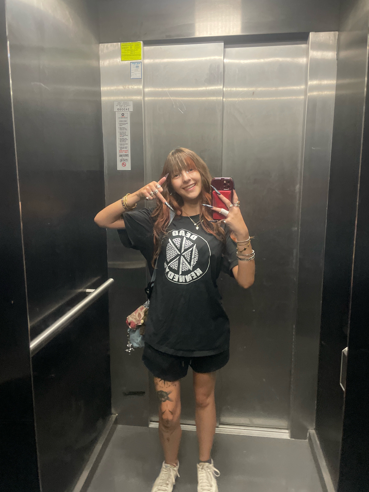 A young woman with long, dark hair with reddish highlights and bangs, wearing a black t-shirt with a circular graphic and black shorts, stands in a stainless steel elevator. She is holding a red smartphone in her right hand, taking a selfie. She is wearing several rings on her fingers and bracelets on her wrists. Her left hand is raised, with her index and pinky fingers extended, forming a peace sign. She has visible tattoos on her left leg and is wearing white sneakers. A small floral-patterned bag is hanging from her shoulder. The elevator has a metal handrail on the left side and visible signs on the wall. The floor is gray. The environment appears to be an elevator in an urban setting, possibly in Raanana, Israel, given the text on the elevator signage which includes Hebrew characters.