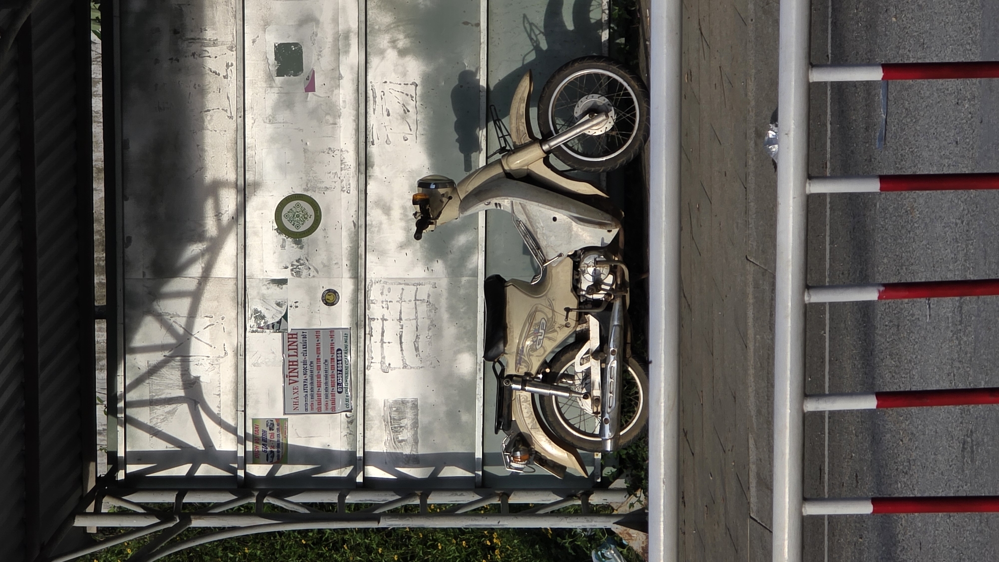 An aerial shot presents a motorcycle parked against a weathered wall in Bo Y, Vietnam. The wall, constructed of white and gray panels, bears graffiti, faded stickers, and a business advertisement in Vietnamese. The advertisement promotes taxi services between Ayun Pa, Ngoc Hoi, and the Po To border crossing. The motorcycle, which is light beige, is parked sideways to the wall, partially visible. To the right of the motorcycle, a barricade with alternating white and red stripes is present, placed alongside a paved surface. The scene is bathed in bright daylight, revealing the wear and tear of the environment. A metal structure, potentially a bridge or overpass, casts a shadow on the wall, and greenery can be seen below it.