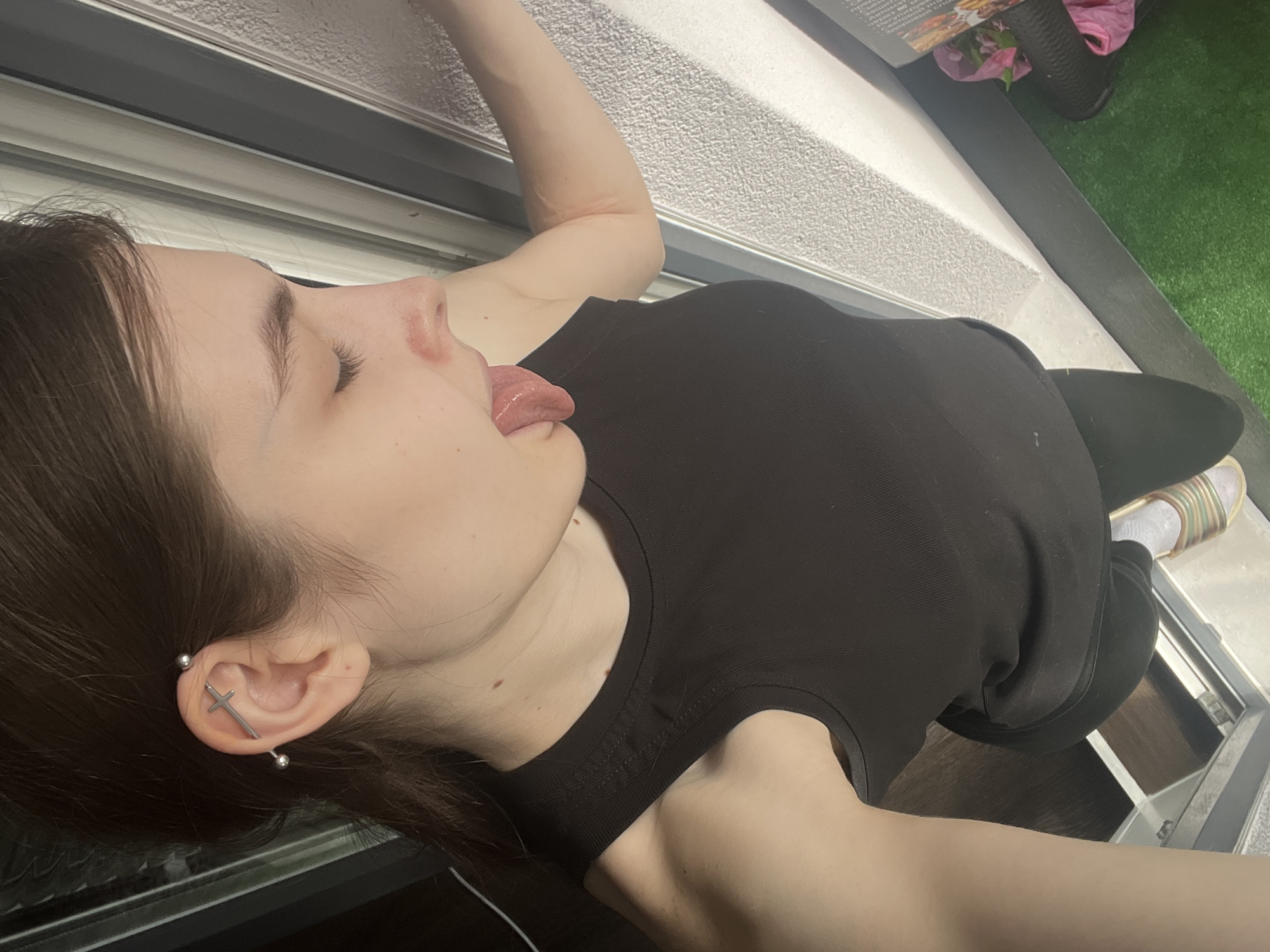 A young woman with brown hair and a cross earring is lying on her back, with her tongue sticking out. She is wearing a black tank top and black leggings, and a white sock and striped sandal are visible on her right foot. The background includes a white wall with a gray window frame, as well as artificial grass. The location in the city of Saint Petersburg, Russia is not discernible from the image.