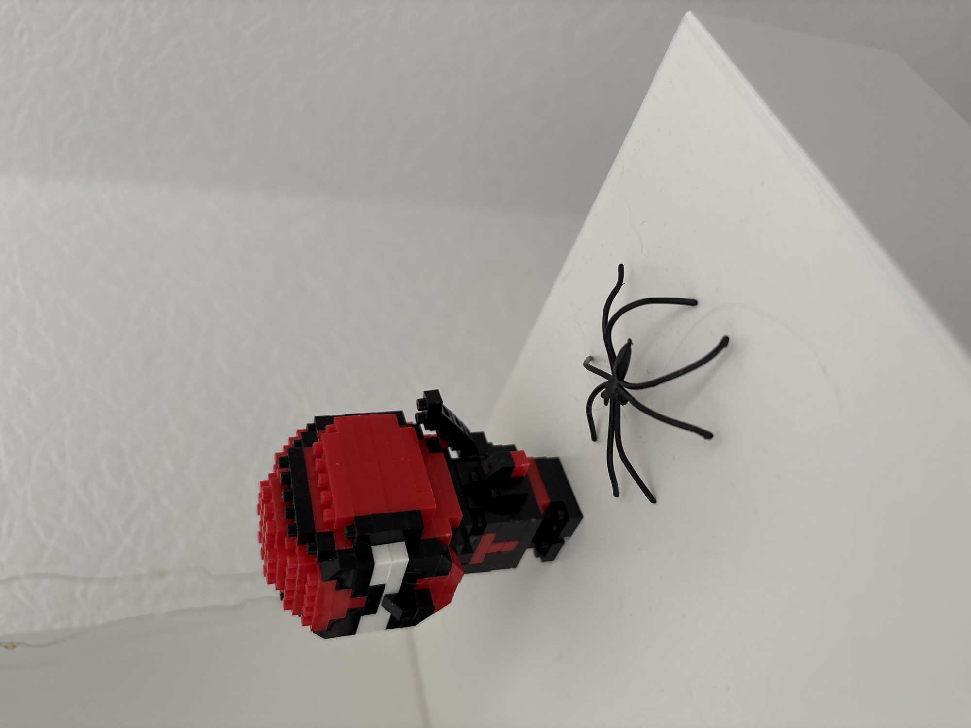 A pixelated red and black figurine, resembling a superhero, stands on a white surface. To its right, a black wire spider with eight legs is positioned on the same surface. The background is a plain, textured white wall. The image is taken from a slightly low angle. There is no indication of the location in Kyiv, Ukraine.