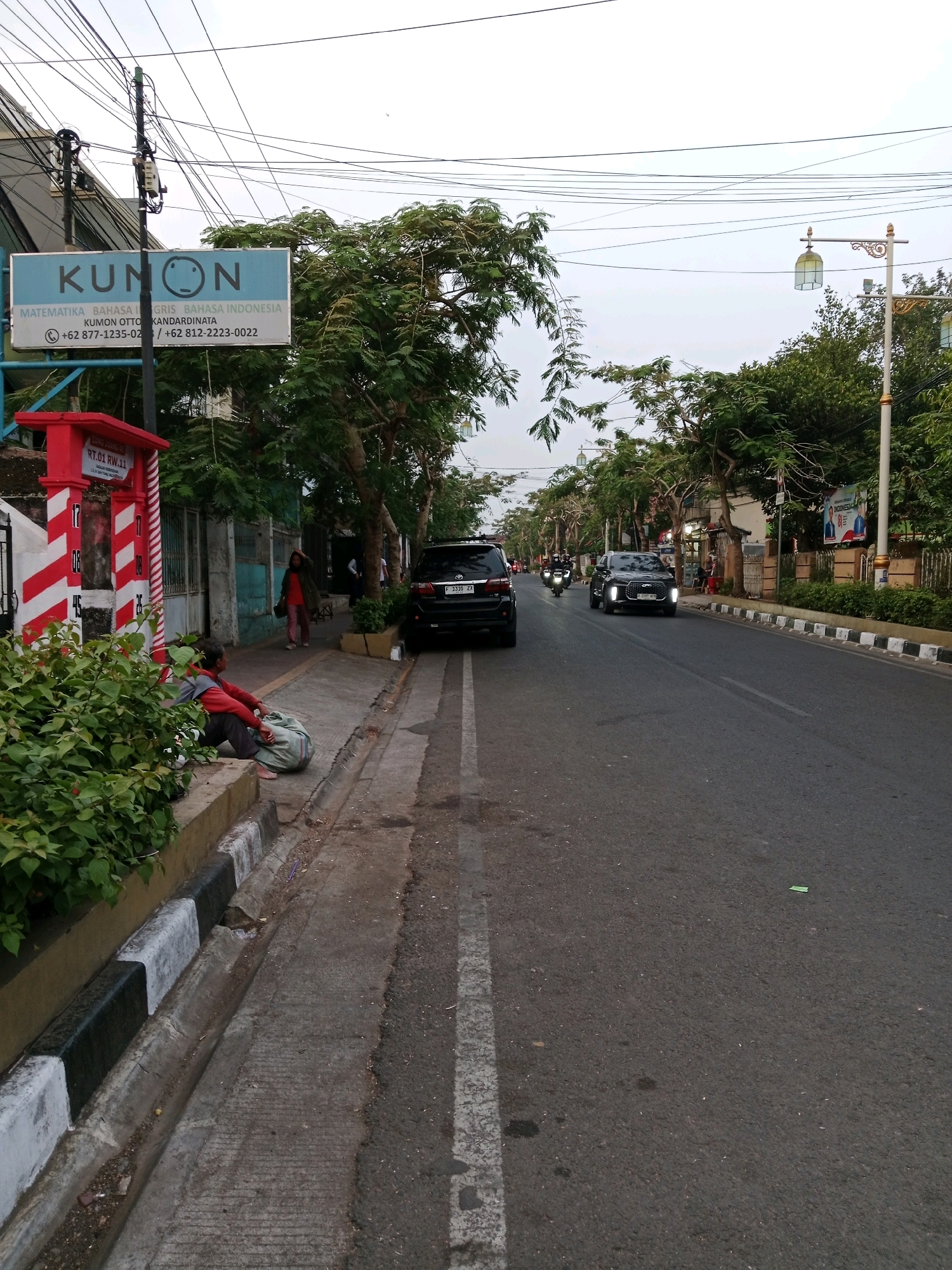 The image depicts a street scene in Cianjur, Indonesia. A sign for "KUNON" is visible on the left, advertising educational services in mathematics, English, and Indonesian. Adjacent to the sign is a red and white striped structure, possibly a public information booth. In the foreground, a person is sitting on the curb, resting a large sack. The street is paved and has a curb with black and white stripes. Trees line the sidewalk, and several vehicles, including cars and motorcycles, are present on the road. A lamppost with an ornamental lantern stands on the right side of the street. The sky is overcast.