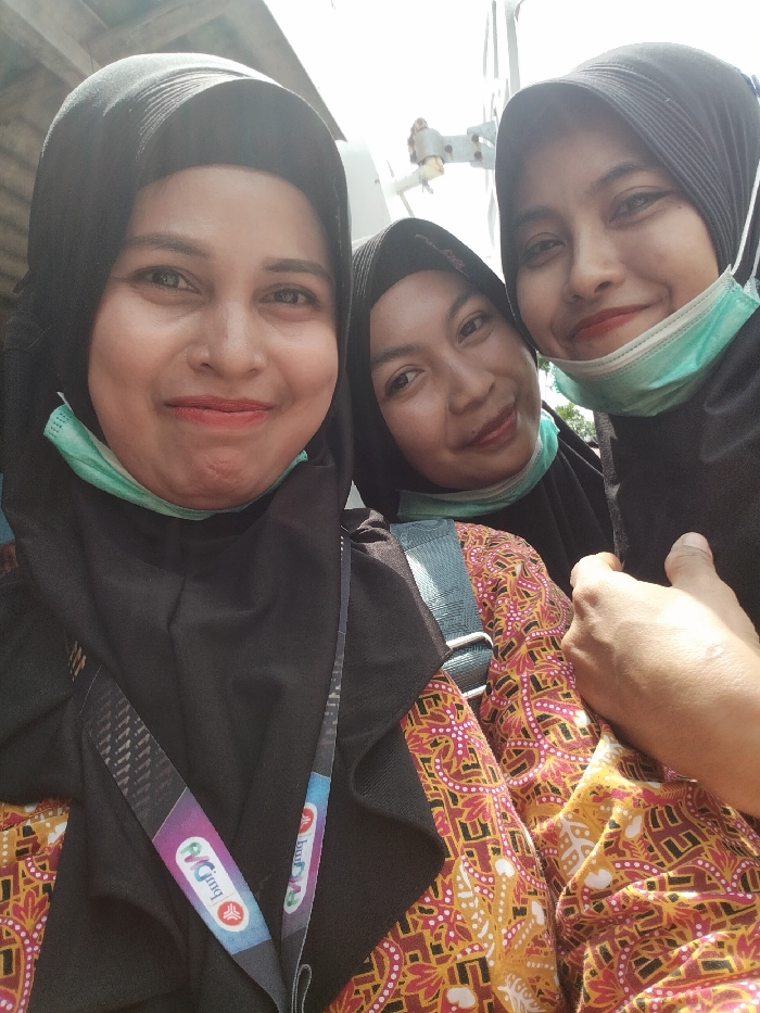 This image depicts three adult females posing closely for a selfie in Kandangjati Kulon, Indonesia. All three individuals are wearing black hijabs and light teal surgical-style face masks, which are pulled down under their chins, exposing their mouths and noses. Their expressions indicate smiles.
The woman on the far left, prominently featured, wears a black top/dress and has a multicolored lanyard with the text "MCJmq" visible across her chest. The woman in the middle, positioned behind and slightly to the right of the first, wears a black top and an orange, yellow, and red patterned batik-style garment, with a blue or grey strap visible over her right shoulder. The woman on the far right, partially visible, wears a black top/dress, and her left hand is raised in the foreground, possibly holding the camera.
The background is brightly lit, primarily white or light grey, indicating an outdoor setting or a well-lit area. Some metallic or structural elements are partially visible in the upper right portion of the background.
