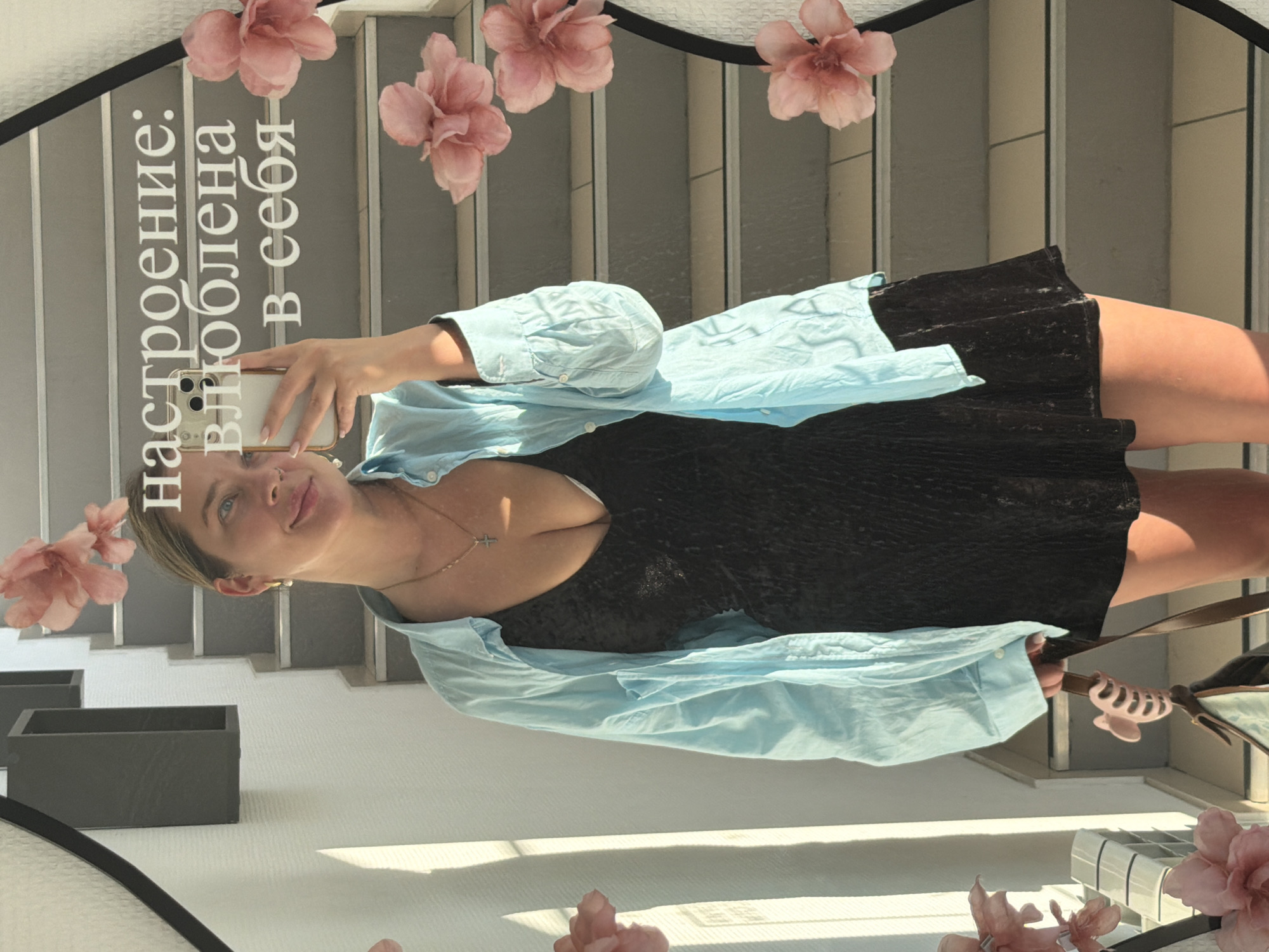 A young woman is captured in a mirror selfie, standing in an indoor setting in Chaikovsky, Russia. She appears to be in her 20s or early 30s, with light hair styled back and a gentle, self-assured smile. Her light blue eyes look towards her reflection as she holds a smartphone to take the picture. She is dressed in a black, possibly velvet, mini-dress, layered with an unbuttoned, light blue long-sleeved shirt. A delicate necklace with a small cross pendant and a subtle nose piercing are visible.
The background reflected in the mirror shows a contemporary interior featuring a staircase with light-colored steps and dark architectural accents. Rectangular dark planters or boxes are visible on a white surface to the left. Bright natural sunlight streams into the space, casting shadows and indicating a daytime scene. Decorative pink flowers, possibly appliqués or stickers, adorn the upper part of the mirror's surface.
Prominently displayed on the mirror, in white Cyrillic script, is the text: "настроение: влюблена в себя". This translates to "Mood: in love with myself," conveying a sense of self-acceptance and happiness that aligns with the woman's serene expression. The overall impression is one of contentment and modern style.