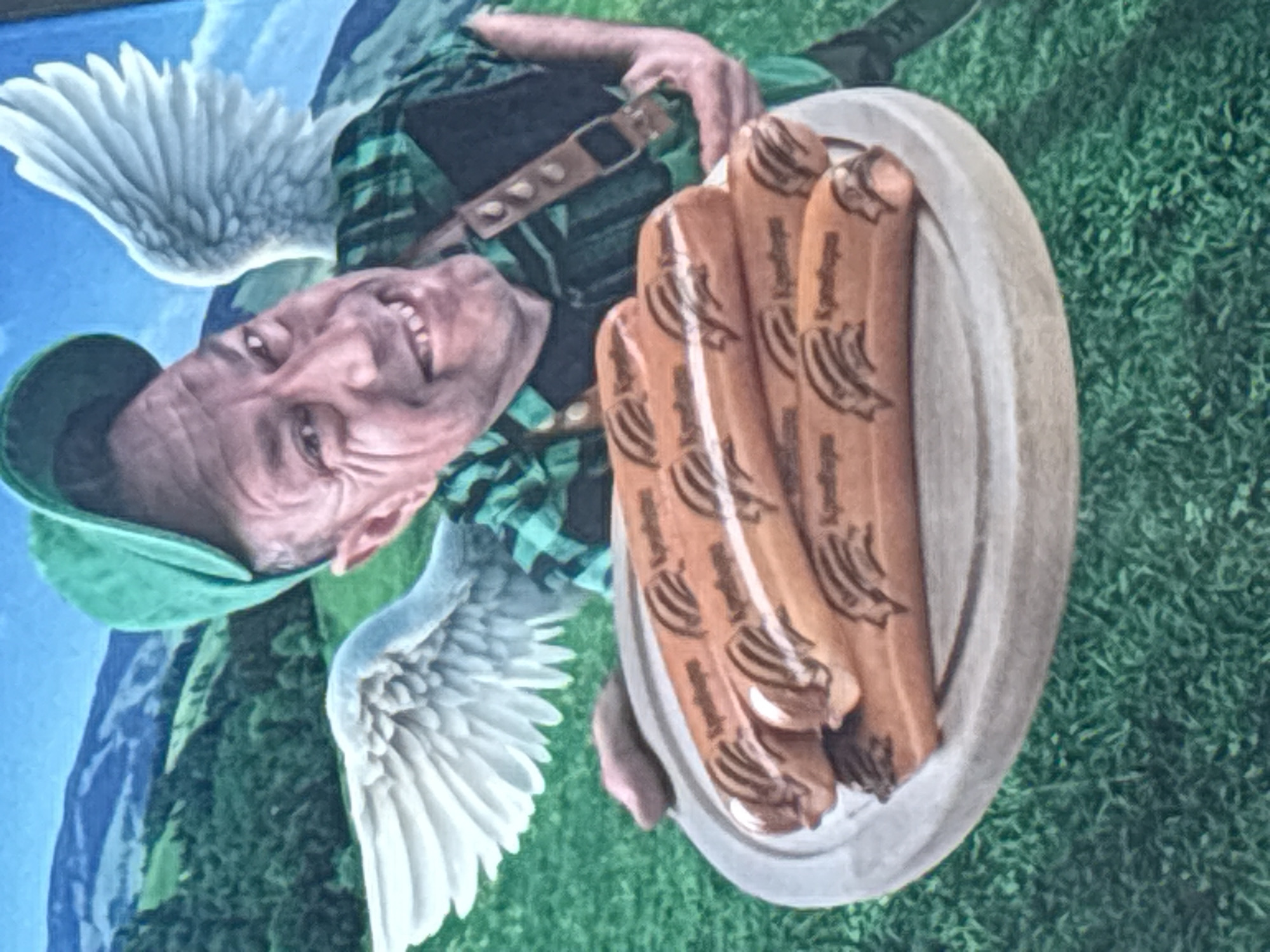 The image appears to be a photograph of a mural or painting, possibly located in Plovdiv, Bulgaria. The scene depicts a man with white wings, smiling, wearing a green cap and a green and black checkered shirt. He also has a dark-colored harness with studs over the shirt. He is holding a plate with four brown sausages, with the word "Kyndinges" inscribed on them, that are placed on a grey plate. The background of the scene consists of a blue sky and snowy mountain peaks on the left side, green grass on the right side and green forest in between. The mural is painted on a rough surface.