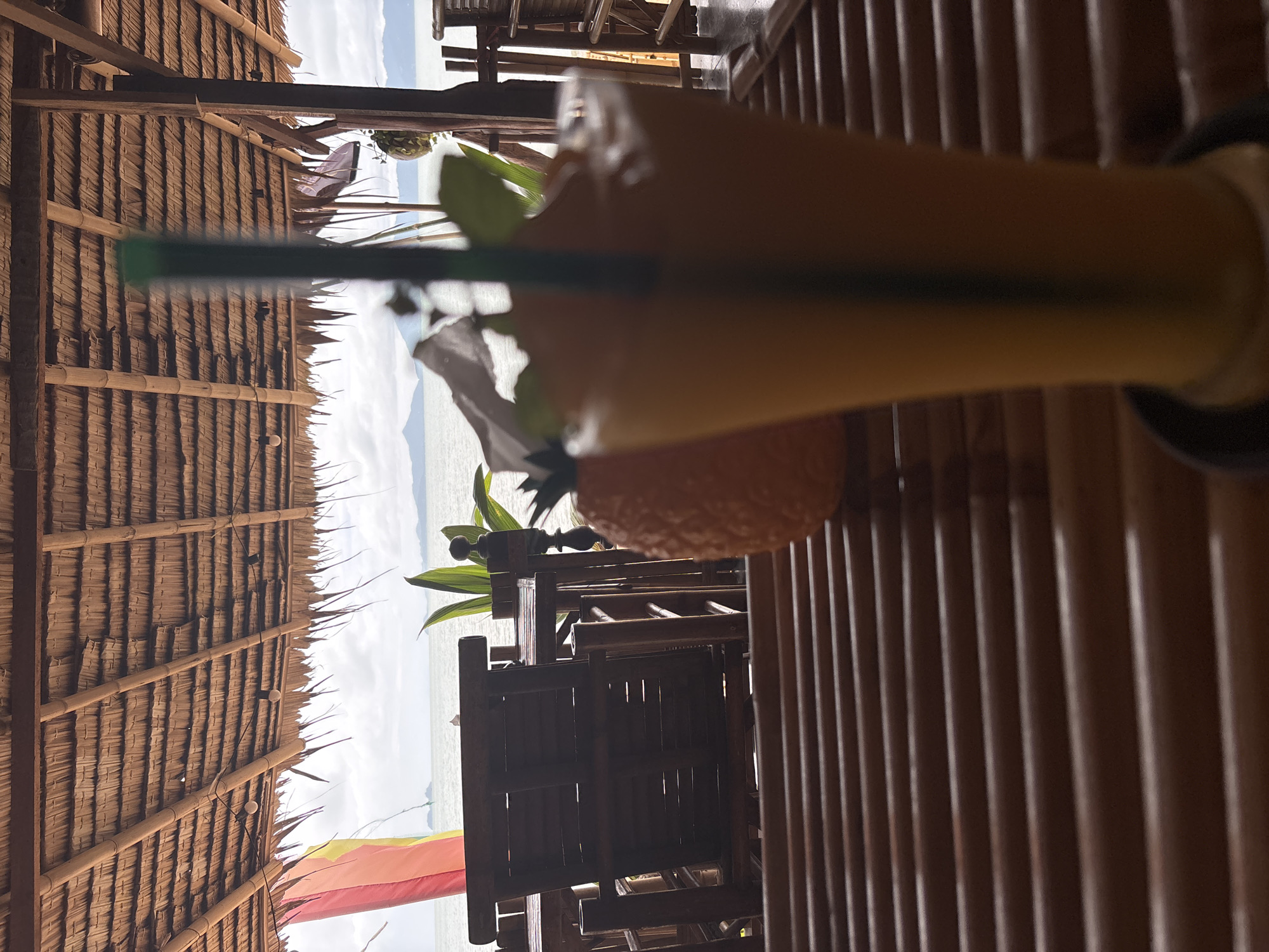 The image shows a close-up, somewhat obscured view from a possible restaurant setting in Baan Bo Phut, Thailand. A tall glass filled with an orange-colored beverage and a green straw dominates the foreground, placed on a dark-colored surface. The environment consists of bamboo structures: a fence to the right with vertical bamboo slats and a thatched roof above made of a darker, likely dried, plant material. A glimpse of a cloudy sky, water, and a distant landmass is visible through the openings. A dark-colored bamboo table and chairs are also present in the mid-ground, as well as a red and yellow flag.