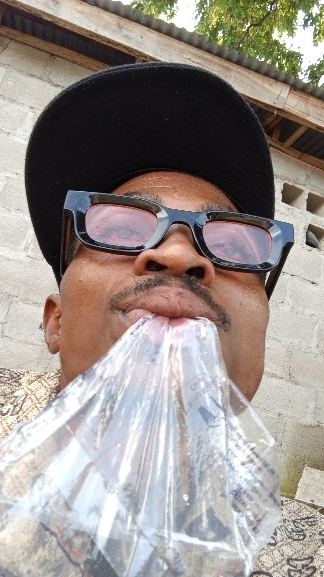 A low-angle, waist-up shot shows a Black man wearing a black baseball cap and black-rimmed glasses. He has a mustache and is holding a clear plastic bag up to his mouth as if to drink from it, or having just drunk from it. The man is wearing a patterned brown and gold shirt. The background consists of a grey brick wall, corrugated metal roofing, and green foliage. The scene appears to be outdoors in the city of Zango-ndaji, Nigeria.