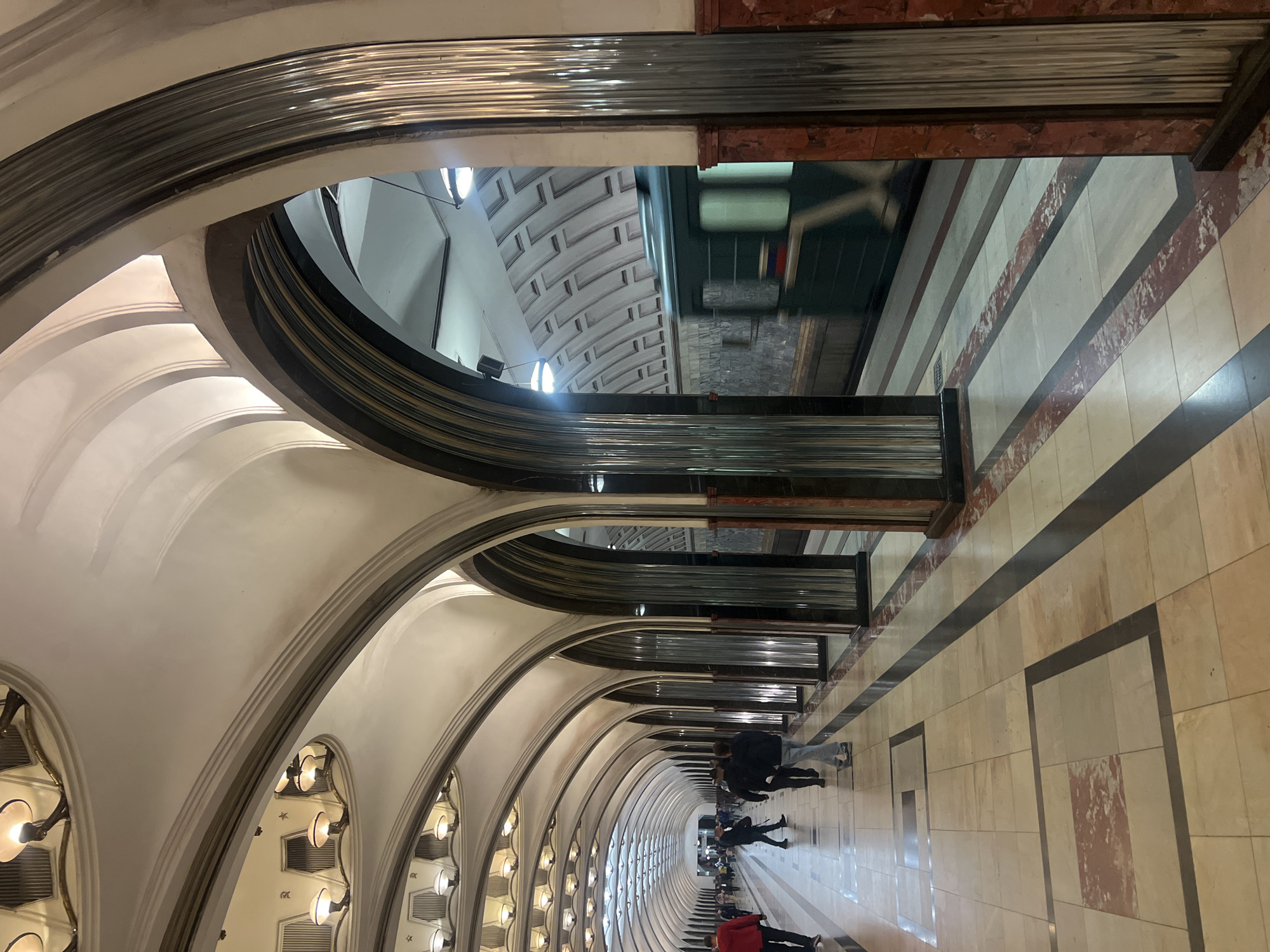 The image shows the interior of a subway station, likely in a large city due to its elaborate architectural details. A series of arches spans the length of the platform, with evenly spaced light fixtures illuminating the vaulted ceiling. Passengers are visible walking along the platform, which is tiled in a light color with darker accent stripes. A subway train is partially visible through an opening above the platform, suggesting it is either arriving or departing from the station. The overall style of the station architecture is reminiscent of classic subway design.