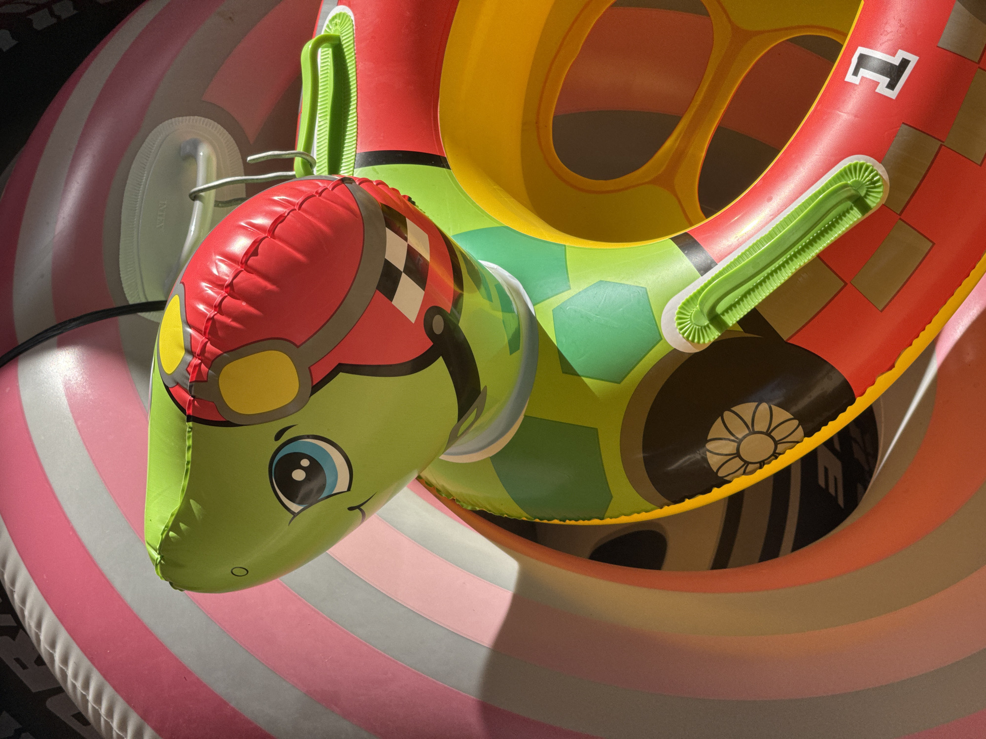 An overhead shot shows several colorful inflatable pool toys. The primary toy is a turtle-shaped float with a red helmet and a cheerful cartoon face. This float is placed atop a larger, striped inflatable ring with bands of pink, white, and grey. Another pool float shaped like a race car is also visible with vibrant colors like red, yellow and green. The image was likely taken outdoors. The city of Kuta Selatan, Indonesia, cannot be verified from the provided image.