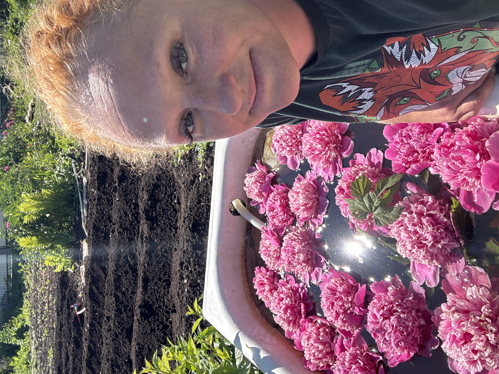 In an outdoor setting, likely a dacha garden in Chaikovsky, Russia, a woman with reddish-blonde hair is captured in a selfie. She is positioned to the right of the frame, her face turned towards the camera with a slight smile. Her eyes are green, and she is wearing dark makeup on her eyelids. The background shows a prepared garden bed with dark soil, and to the left, a large bathtub filled with water and numerous bright pink peonies floating on the surface. A white hose is visible emerging from the bathtub's edge. The sunlight creates a lens flare in the image. The woman is wearing a dark green t-shirt with a colorful, stylized fox graphic on the sleeve. In the background, there are green plants and a metal fence.