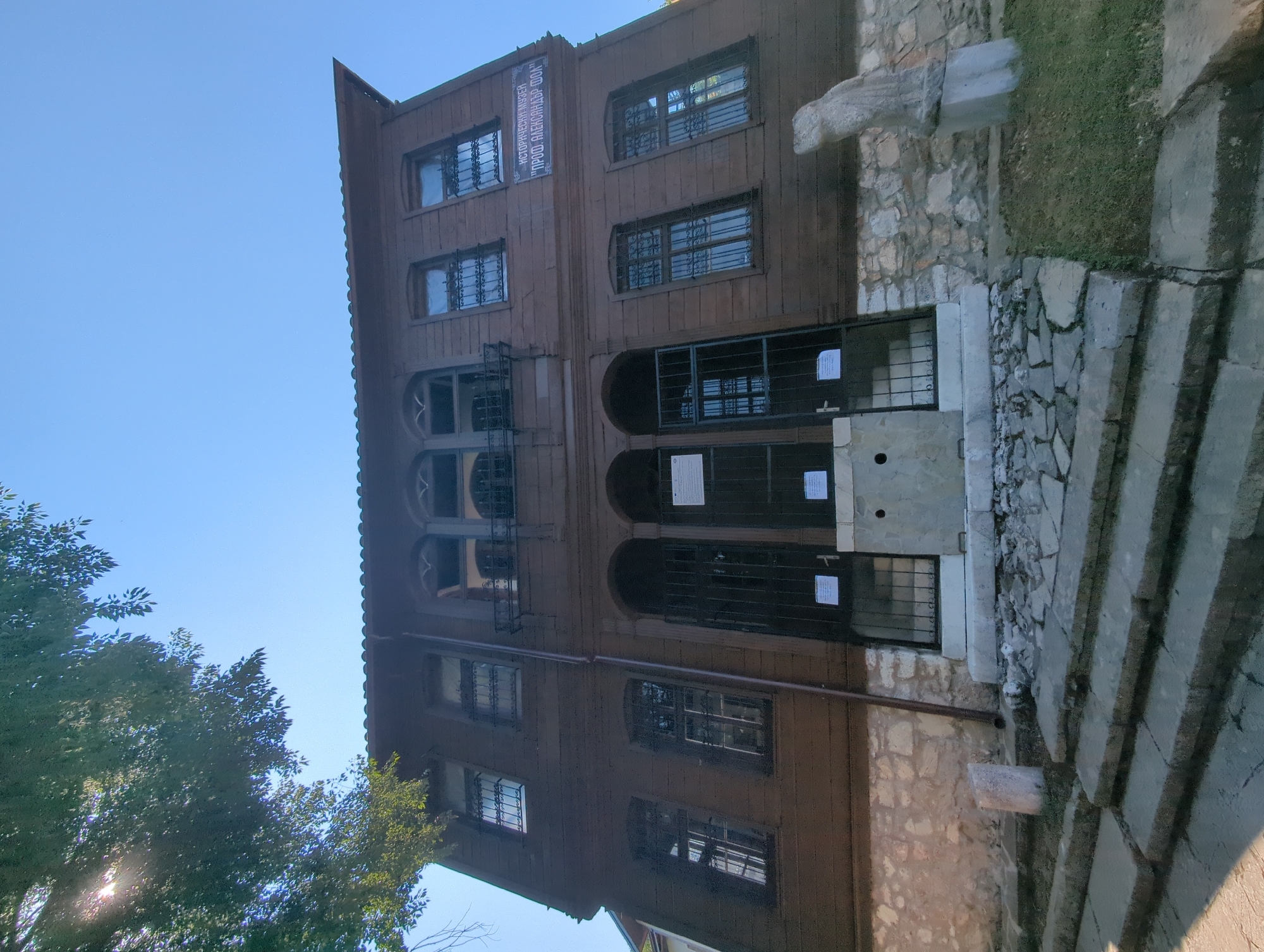 The image shows the facade of the Historical Museum "Prof. Aleksandar Foll" in Malko Tarnovo, Bulgaria. The building is a two-story wooden structure with stone foundations. The first floor has three arched entrances, each covered by a black metal gate. The second floor has several windows, some with arched tops and black metal grilles. A sign above a window on the right side of the second floor reads "ИСТОРИЧЕСКИ МУЗЕЙ "ПРОФ. АЛЕКСАНДЪР ФОЛ"". In front of the building, there are stone steps leading up to the entrance. To the right of the steps, a stone statue of a reclining figure is visible on a pedestal. The sky is clear and blue, and a large tree casts a shadow over the top left corner of the building. The overall impression is that of an old, well-preserved historical building.