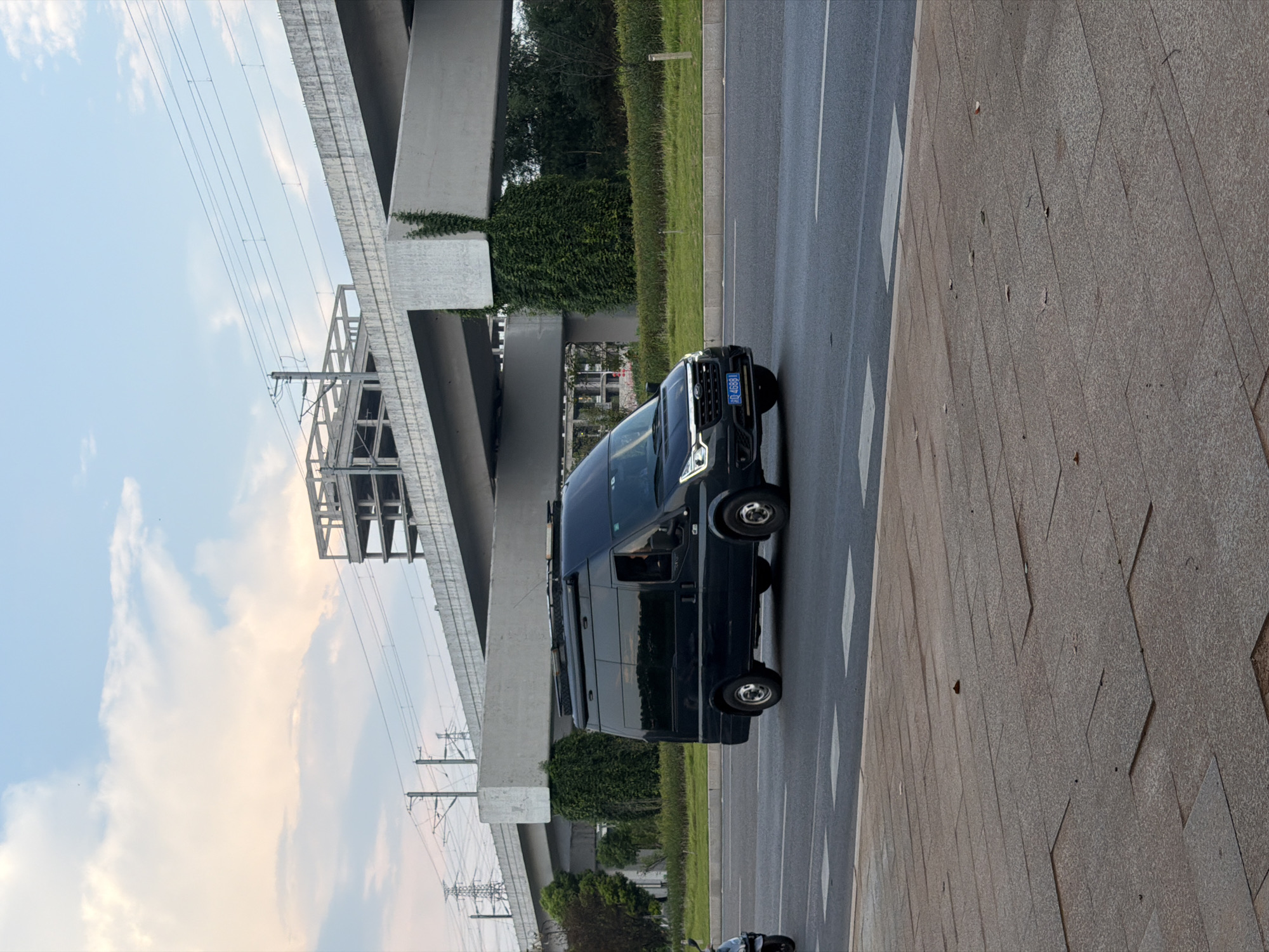 A black van is driving on a road in Jiangjin, China. The van has a license plate that reads "Q 46881". In the background, there is a large concrete overpass with train tracks running on top of it. There are also some power lines and a partially constructed building in the distance. The sky is partly cloudy with a sunset glow. The road has white lane markings and a pedestrian crosswalk. The foreground shows a paved sidewalk with large, textured tiles. A portion of a motorcycle is visible on the left side of the image, partially obscured by the van.