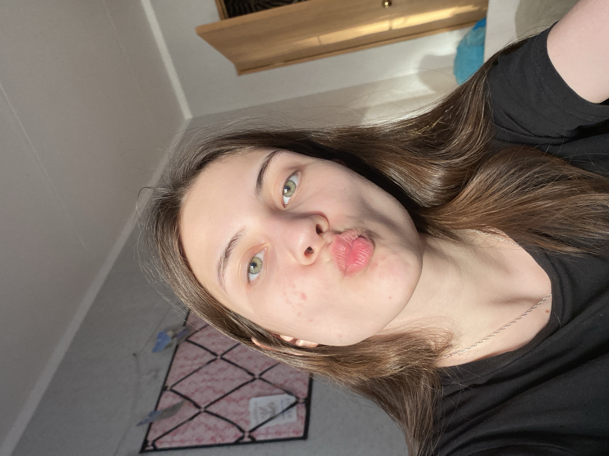 A young woman with fair skin, green eyes, and light brown hair is posing for a selfie. Her lips are pursed in a kissing gesture. She is wearing a black shirt and a delicate silver necklace. The background includes a light-colored wall, a piece of furniture with a light wood finish, and what appears to be a framed board with a grid pattern and pink accents. The location is potentially within a residential space, but the city of Votkinsk, Russia, cannot be definitively confirmed based on the visual information alone.
