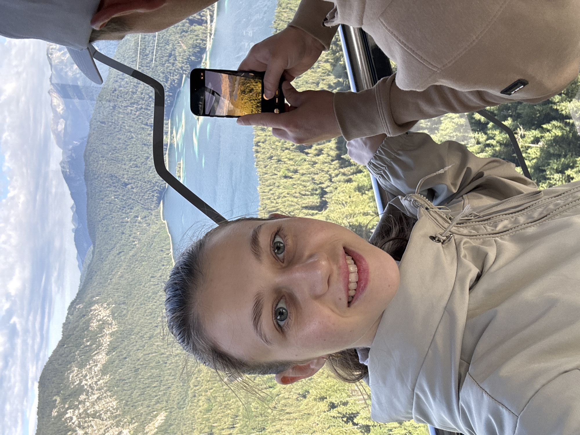 In Grainau, Germany, a young woman with light brown hair pulled back and blue eyes smiles at the camera, her head and shoulders filling the foreground of the image. She is wearing a light beige hooded jacket. To her right, a person wearing a beige hoodie and a light gray baseball cap is holding a smartphone, taking a photograph of the landscape. The phone screen displays a scenic view of a lake surrounded by a dense forest of green trees, with mountains in the background. The woman's gaze is directed towards the viewer, while the person with the phone is looking at the phone screen. The backdrop is a panoramic view of a vast mountain range with a large, blue lake nestled within a valley. The sky is partly cloudy with patches of blue. The scene appears to be captured from an elevated perspective, possibly from a cable car or a high vantage point, overlooking the Eibsee lake and the surrounding Zugspitze massif.