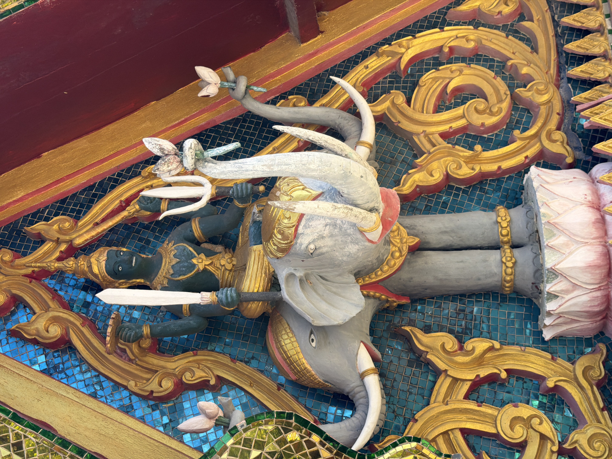 The image shows a close-up of a detailed stucco sculpture, likely part of a temple or shrine decoration, located in Baan Plai Laem, Thailand. The sculpture depicts a multi-armed deity with dark skin, wearing a golden headdress and ornate jewelry. The deity is seated on the back of a large, gray elephant with prominent tusks. The elephant is adorned with golden trappings, including a headdress and a decorative collar. The deity holds a white sword in its right hand and a trident in its left. The background is a mosaic of small, turquoise tiles, framed by elaborate golden and red ornamental carvings. Two pink lotus flowers are depicted, one held by the deity's left hand and another near the elephant's trunk, also appearing to be held by a separate, smaller elephant trunk-like appendage. The overall scene is colorful and highly decorative, characteristic of Thai Buddhist art.