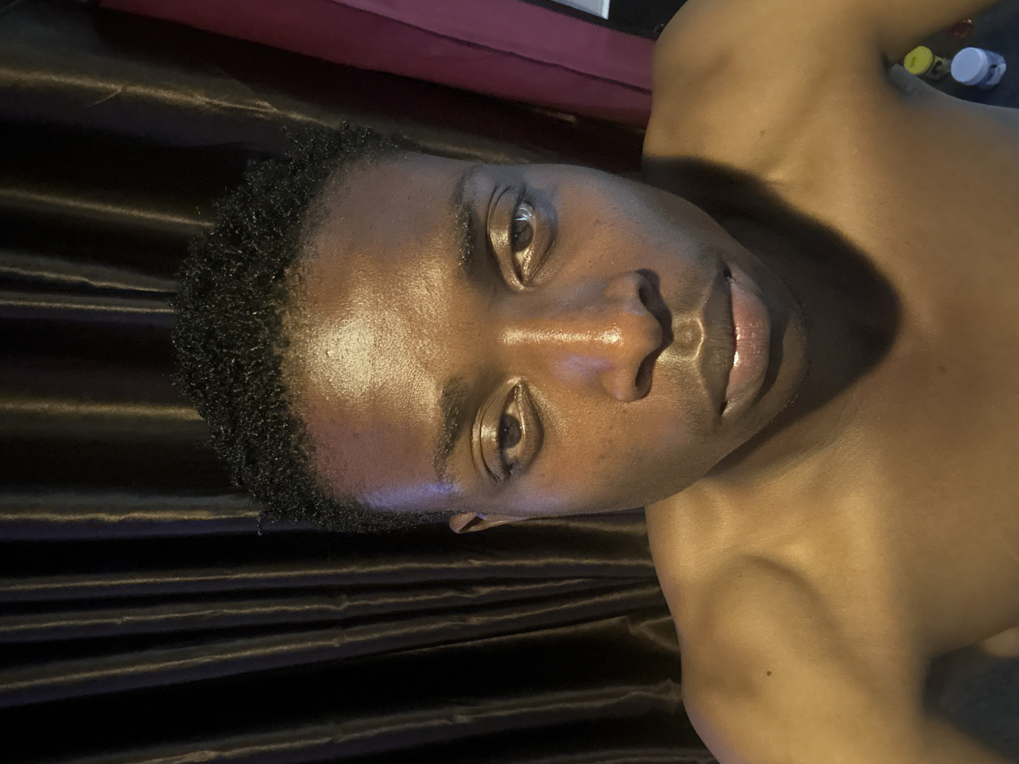 A medium, close-up, slightly angled shot depicts a dark-skinned, shirtless person, presumably a male, with short, curly black hair. The person's face is well-lit, highlighting features such as the nose, eyes, and lips. The background includes a dark, textured surface that resembles draped fabric, and a glimpse of what appears to be a red object in the top portion of the image. The photograph does not provide enough context to confirm a specific location within Bukuru, Nigeria. The overall scene suggests a personal or casual setting.