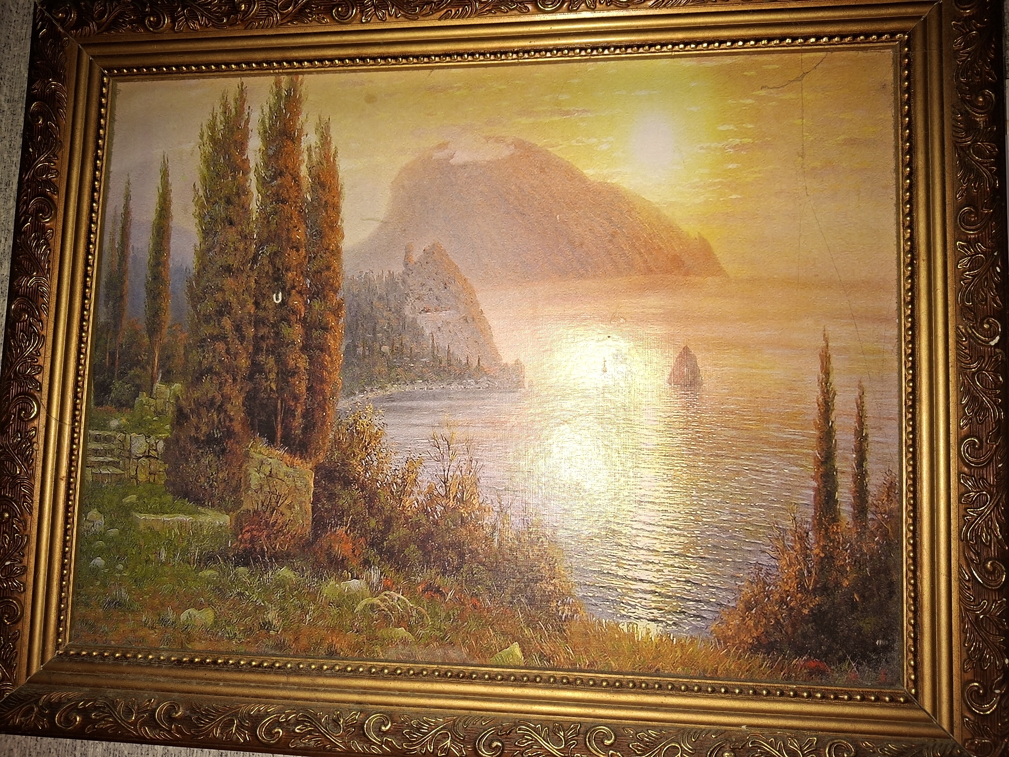 This is a framed oil painting depicting a coastal landscape at sunset. The painting shows a body of water reflecting the warm, golden light of the setting sun. In the foreground, a grassy, rocky shoreline is visible, dotted with trees and shrubs. Several tall, slender cypress trees are prominent on the left side of the painting. In the middle ground, a mountainous landmass rises from the water, with a small settlement or fortress built on its slopes. The sky is a gradient of yellow, orange, and pale blue, indicating the sun is low on the horizon. A small boat is visible on the water in the distance. The frame is ornate and appears to be made of dark wood with gold detailing. The image does not depict any specific identifiable location within the city of Nikolaevka, Russia, or any people.