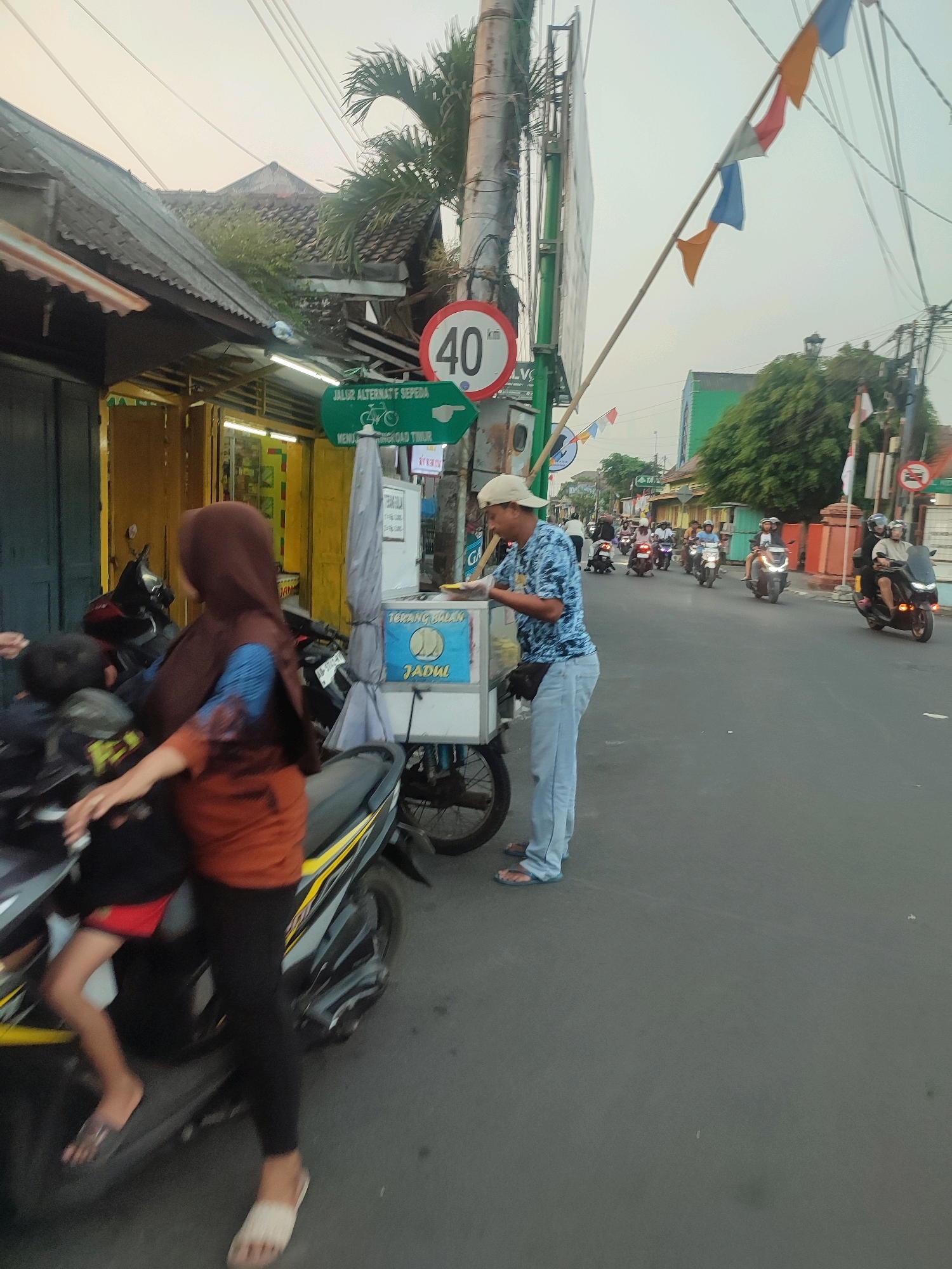 The image captures a street scene in Yogyakarta, Indonesia, likely during the late afternoon or early evening, with soft, diffused lighting. A man wearing a light blue patterned shirt, light blue jeans, and a beige cap stands behind a food cart labeled "Terang Bulan Jadul". He appears to be serving or preparing food. In the foreground, a woman wearing a brown hijab and an orange top, along with a child wearing a red and black garment, are seated on a black motorcycle.
The street is paved, and on the right side, several motorcycles are seen traveling in the same direction. The buildings on the left side of the street appear to be shops with open doorways and awnings. A green directional sign indicating "JALUR ALTERNATIF SEPEDA" (Bicycle Alternative Route) with a bicycle icon and an arrow pointing to the right is visible. A traffic sign with "40" indicating a speed limit is also present. Festive flags are strung across utility wires in the background. The overall atmosphere suggests a typical urban environment in Indonesia with moderate activity.