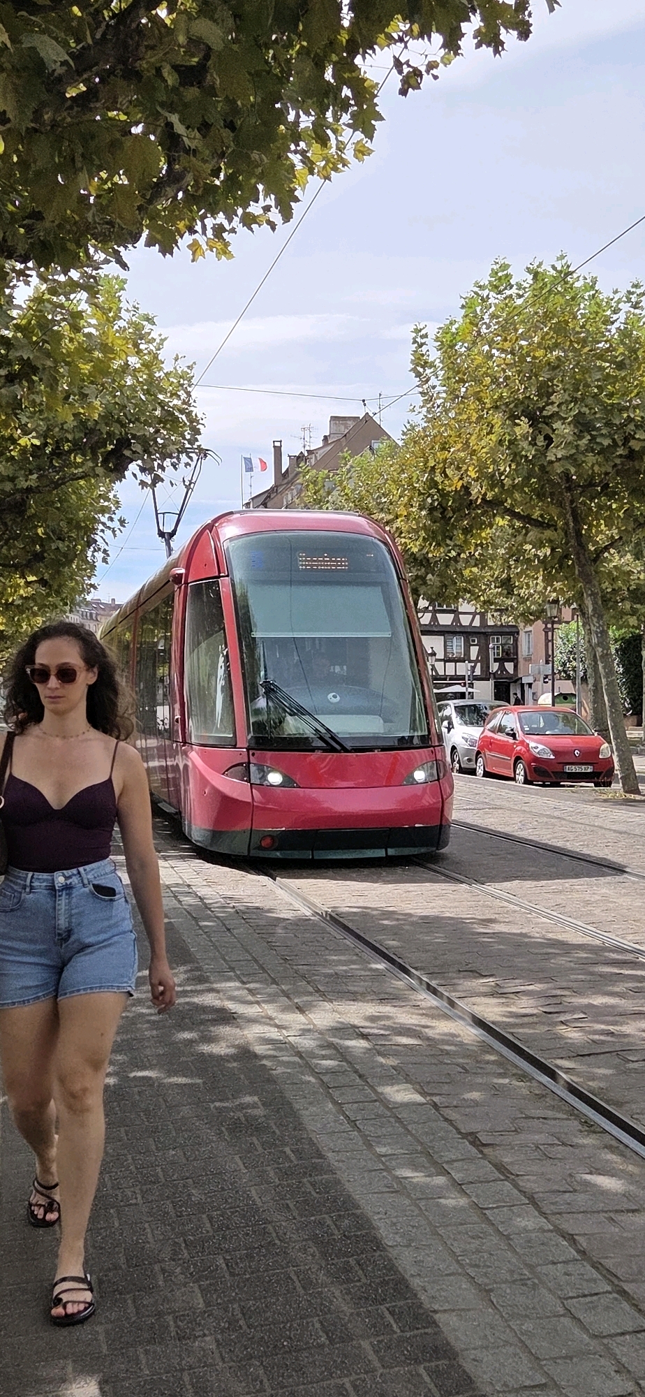 A woman walks on a cobblestone street in Strasbourg, France, on a sunny day. She is wearing a purple top, denim shorts, sunglasses, and sandals. A red tram is passing by her on the tram tracks. In the background, there are trees and buildings. One of the buildings has a distinctive half-timbered facade, characteristic of Alsace. Two French flags are visible flying from a pole. A red car is parked on the right side of the street. The tram has "HOENHEIM" written on its digital display.