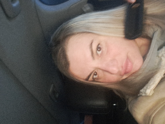 A woman with blonde hair, light complexion, and brown eyes is looking at the camera. She is holding a black hairbrush near her face. She is wearing a tan jacket with a white, fuzzy collar. The background appears to be the interior of a car. The image is slightly blurry and rotated.
