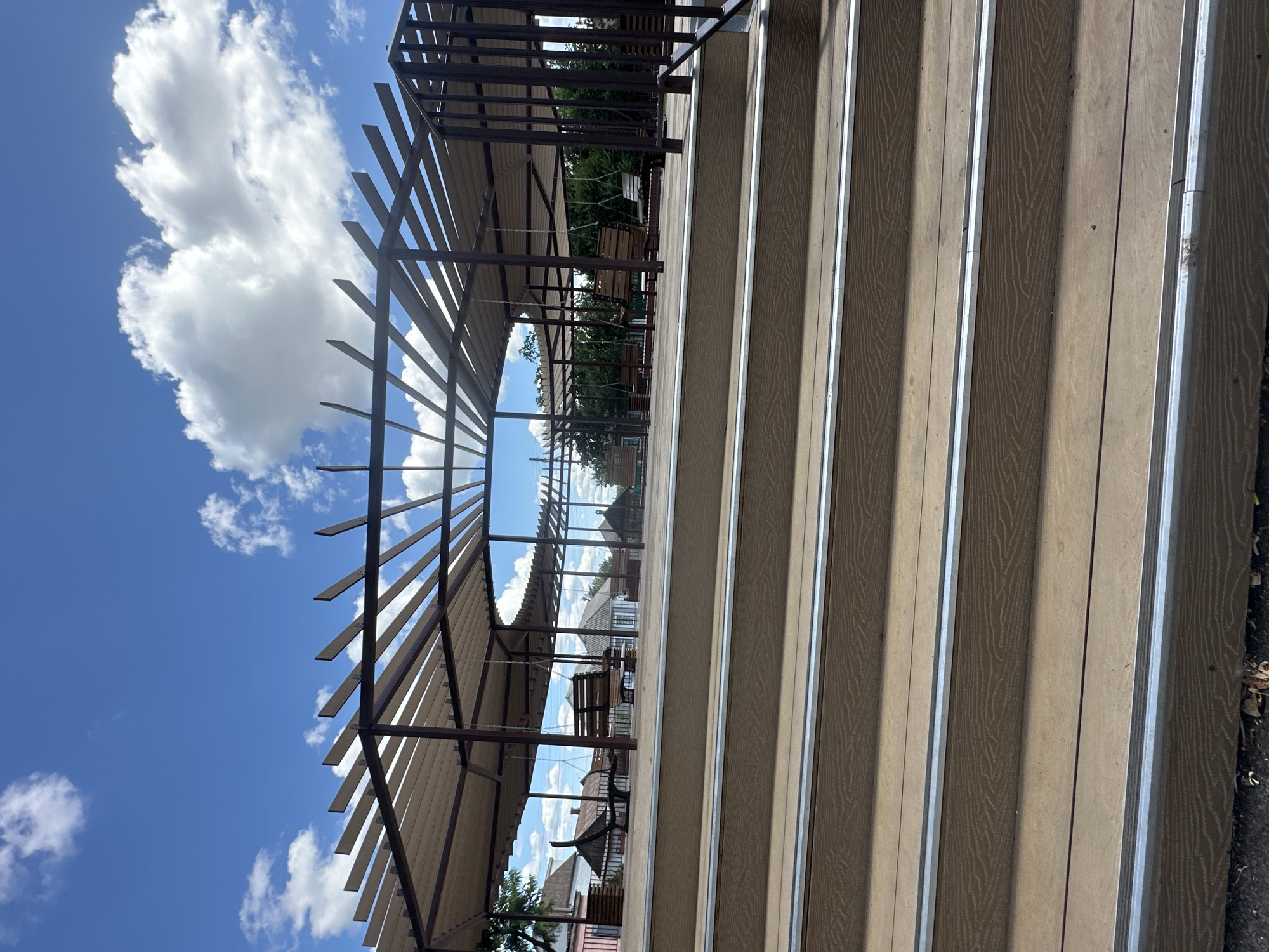 The image depicts an outdoor seating area with benches and swings situated under a covered pergola. The area is accessed by a wide set of wooden stairs with metal trim on each step. The pergola is constructed of dark metal beams and supports, with a light-colored fabric covering the top. Several wooden benches and swings are visible throughout the covered area. In the background, there are buildings with pitched roofs, trees, and a bright blue sky with scattered white clouds. The scene appears to be in a public space, possibly a park or recreational area, in Zhovte, Ukraine. There are no people visible in the image.