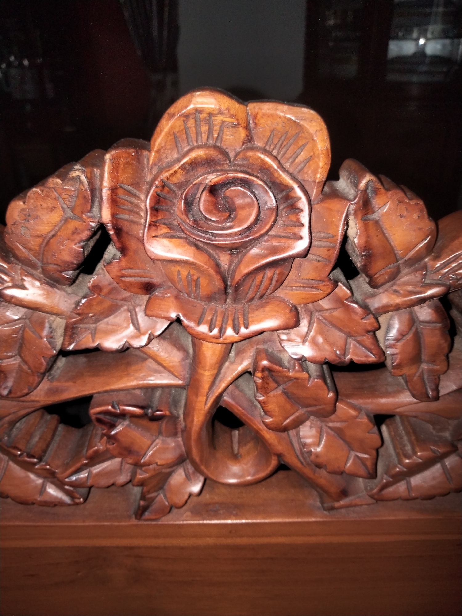 A close-up, slightly angled, eye-level shot shows a dark brown wooden carving of a rose with surrounding leaves and stems. The carving appears to be part of a larger wooden furniture piece, possibly a headboard or decorative panel. The wood grain is visible, and the carving features deep indentations and textured surfaces that create a sense of depth. The lighting is dim, casting shadows that accentuate the three-dimensional quality of the carving. The background is out of focus and appears to be a dark, reflective surface, suggesting the carving is indoors in a dimly lit room. There are no people or animals visible in the image. The specific location of the carving within the city of Payagaboh, Indonesia, cannot be determined from the visual information provided.