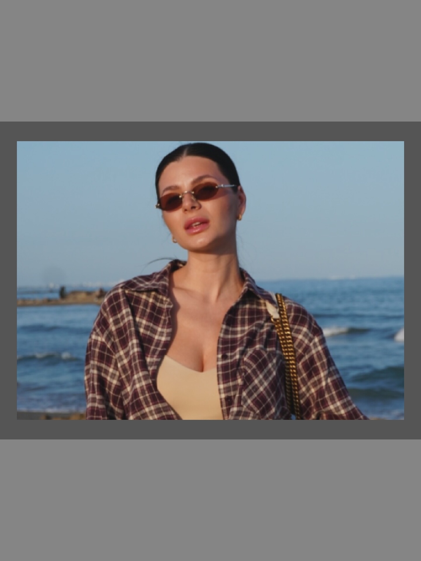 A woman stands on a beach in front of the ocean. She has dark hair pulled back and is wearing sunglasses, a beige top, and a plaid shirt. A gold chain purse is slung over her shoulder. The ocean is blue and calm, with a distant shoreline visible.