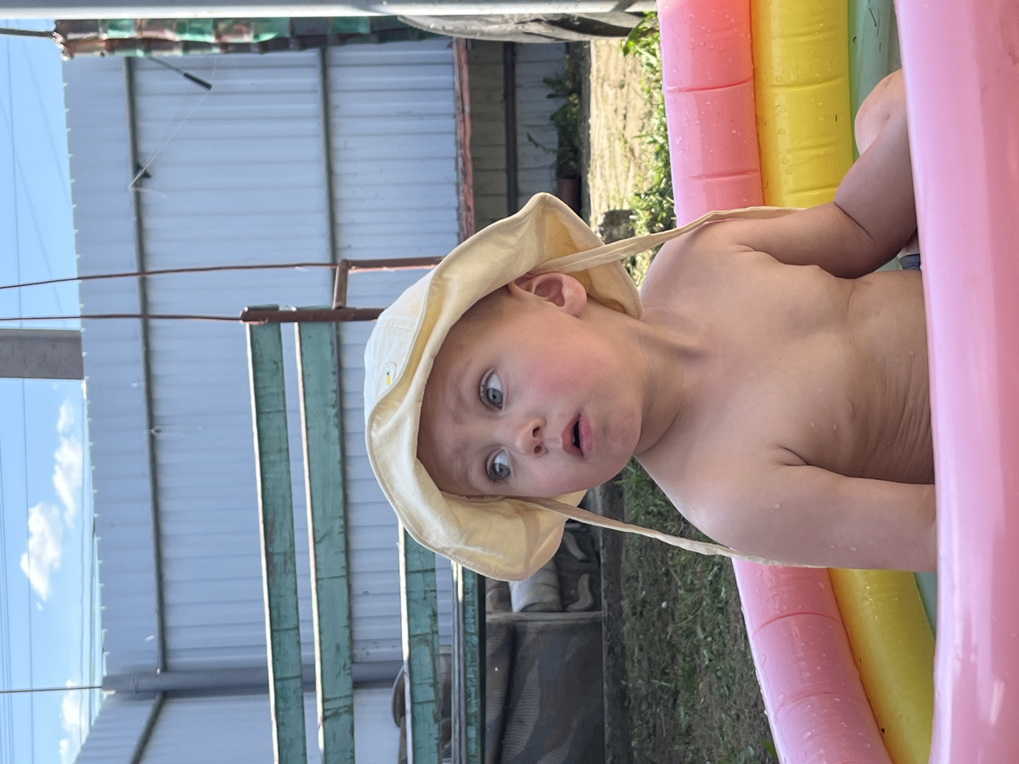 A young child wearing a yellow sun hat is partially submerged in an inflatable pool with pink, yellow, and green sections. The child has a surprised expression. The location appears to be outdoors, potentially in a backyard, with a corrugated metal fence or building in the background. Vegetation and some structures are visible behind the fence. The image was taken in the city of Фоки, Russia.