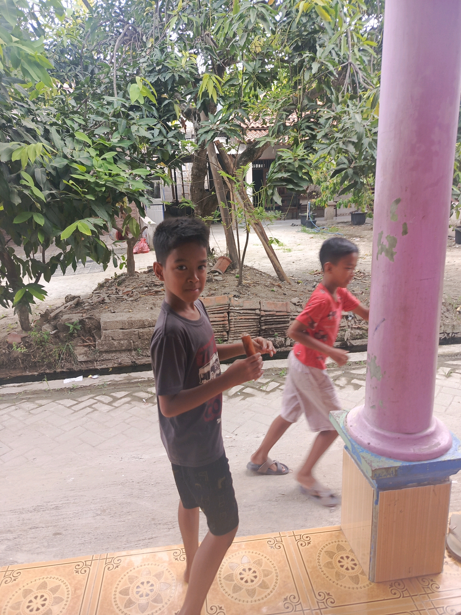 The image captures an outdoor scene in Balaraja, Indonesia, with two young boys as the central subjects. On the left, a boy wearing a dark t-shirt and black shorts stands facing the camera, holding a small orange object in his right hand. To his right, another boy in a red t-shirt and light-colored shorts is blurred as he moves past, heading towards a large, light purple cylindrical pillar. The pillar is partially visible on the right side of the frame and stands on a decorative base.
The environment appears to be a residential area. Lush green trees with dense foliage dominate the background, their branches creating a canopy overhead. In the mid-ground, there are elements of construction or landscaping, including piles of bricks, a small ditch or water channel, and various plants. The ground is paved with light-colored bricks in a patterned arrangement, leading to an area with decorative tiles in the foreground. The overall lighting suggests an overcast or bright daylight setting.