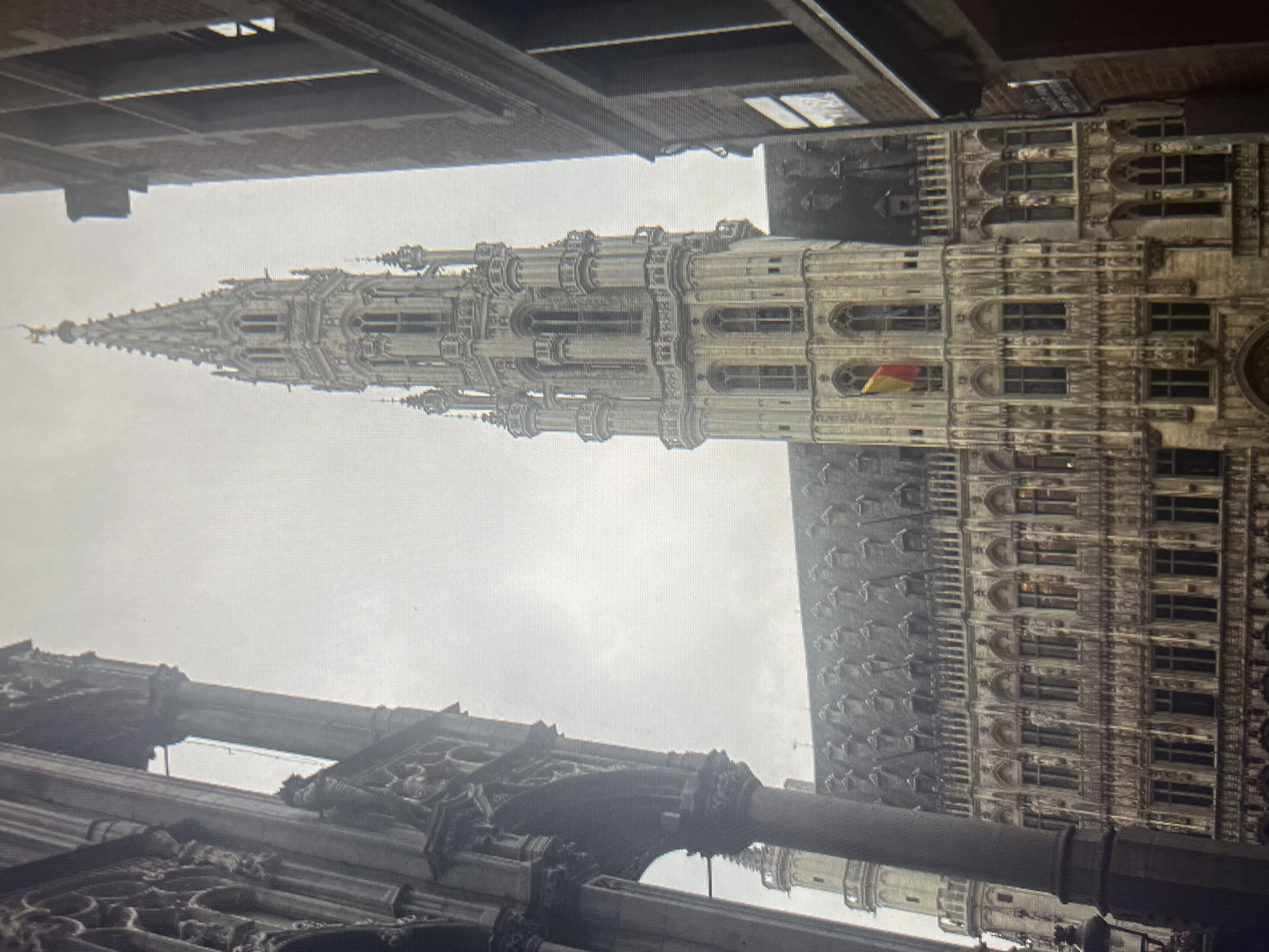 The image shows a view of a tall, ornate building with a spire, likely a historic town hall or cathedral, surrounded by other buildings. The architecture suggests a European city with Gothic or Flemish Renaissance influences. The sky is overcast, casting a dim light on the scene. A flag, appearing to be red, yellow, and black, is visible on the facade of the main building. The foreground is partially obscured by the corner of another building on the left. The scene is devoid of people. The reference to "Qarağandı city administration, Kazakhstan" in the prompt is incongruous with the visual evidence of the architecture.