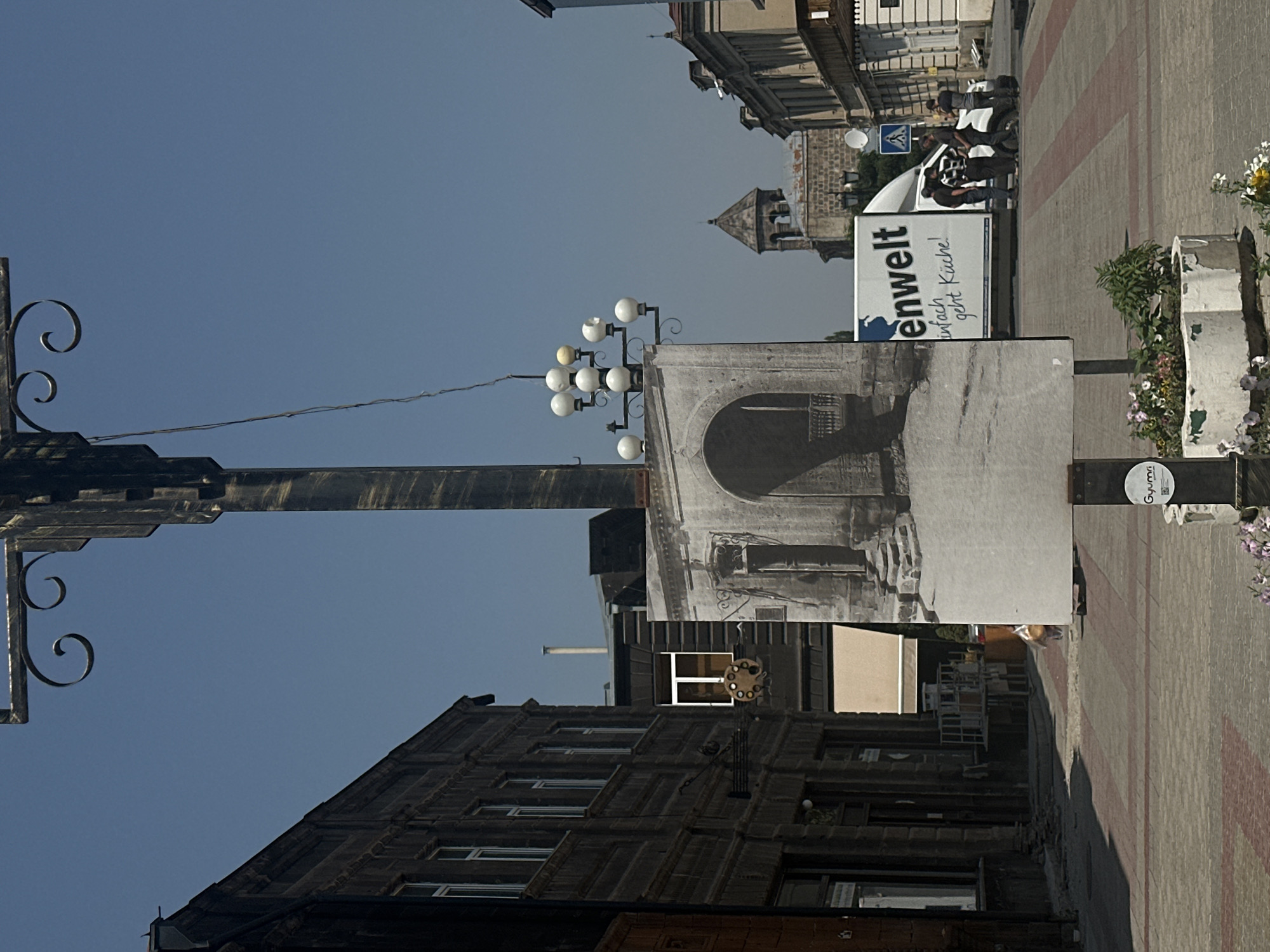 The image depicts an outdoor scene in the city of Gyumri, Armenia, likely on a sunny day. A large, black lamppost with decorative metal scrollwork and multiple light fixtures dominates the center of the frame. Attached to the lamppost is a large, black-and-white photograph or poster featuring an arched doorway with stone steps and classical architectural details.
To the left of the lamppost is a dark-colored, multi-story building with numerous windows. Further down the street, another building with a white-framed window is visible. In the background, on the right side of the image, a white delivery truck with German text ("enWelt" and "einfach geht Küche!") is parked. Several individuals, dressed casually, are gathered around the truck, appearing to be engaged in conversation or unloading. Beyond the truck, a stone tower with a pointed roof and arched windows is visible, indicative of older architecture. The street is paved with red and grey bricks arranged in a patterned walkway. A concrete planter with flowers is situated in the foreground, near the base of the lamppost. The sky is clear and blue.