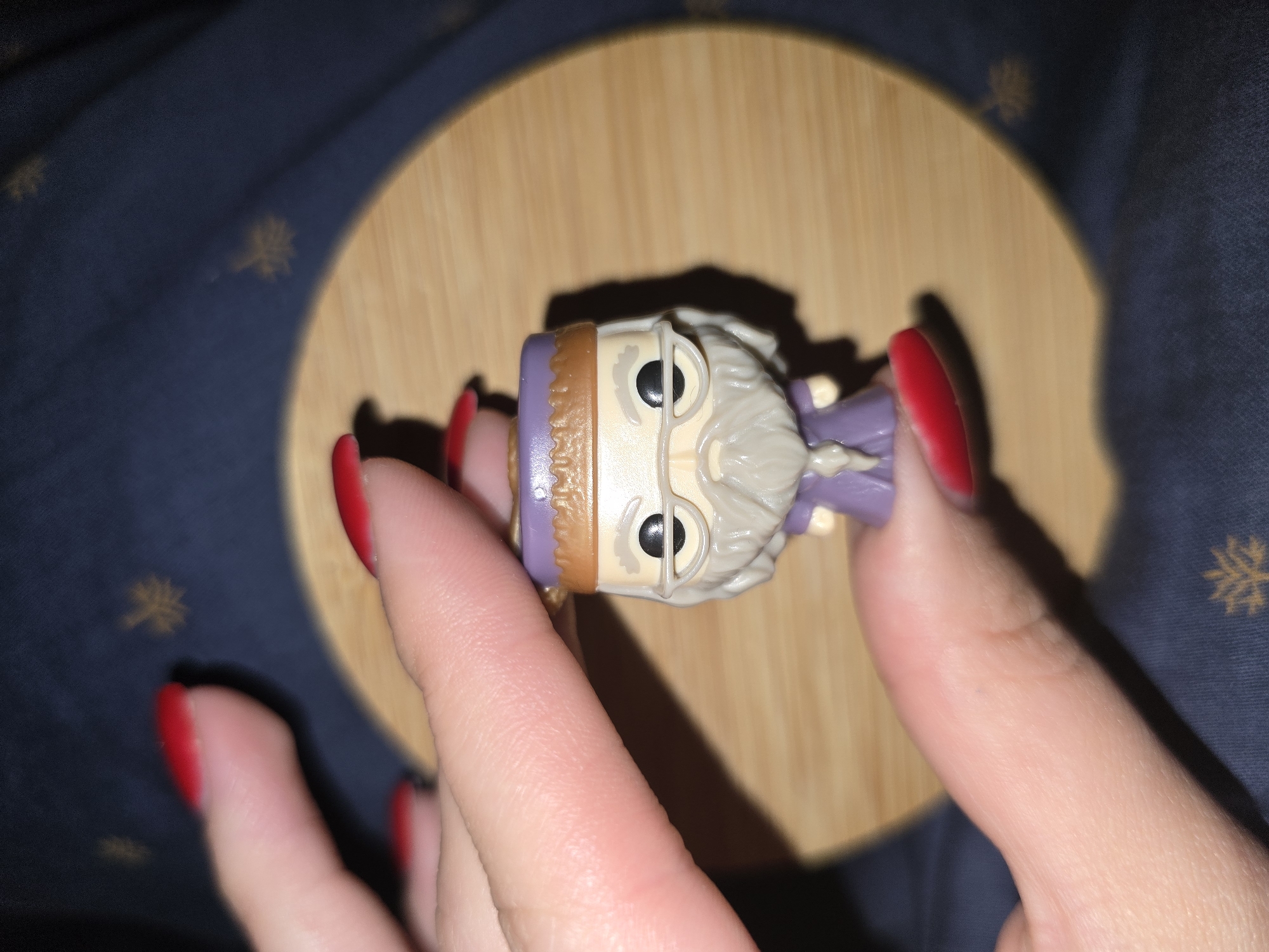 A close-up, overhead shot shows a small figurine of an elderly woman with gray hair, glasses, and a purple robe. She is being held between two fingers of a person with bright red nail polish. The figurine is positioned on a round, wooden surface, which is in turn placed on a dark blue fabric with a subtle gold snowflake pattern. The lighting creates a soft shadow to the right of the figurine. The setting appears to be indoors, possibly a home environment in Petropavlivska Borshchahivka, Ukraine. There is no visible text in the image.