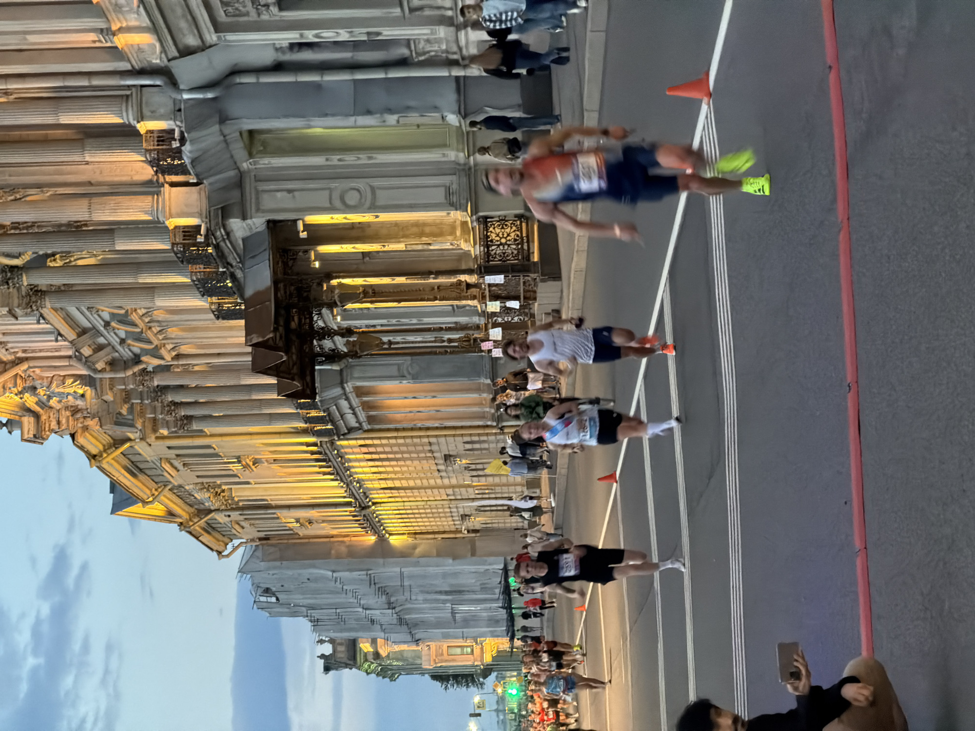 The image depicts a marathon race in Saint Petersburg, Russia, likely near the Hermitage Museum, given the neoclassical architecture of the buildings lining the street. In the foreground, several runners are in motion, their legs blurred by speed. A man in a black tank top and shorts is in focus, wearing bib number 2591. To his right, a woman in a white and blue tank top and black shorts, with bib number 1262, is also running. Further ahead, a runner in a red, white, and blue tank top and dark shorts, with a blurred bib number, is sprinting. Spectators are visible on the sidewalks and in the background, some of whom are taking pictures with their phones. The street is marked with white racing lines and orange cones. The buildings feature grand columns, ornate facades, and balconies, illuminated by warm, artificial light, suggesting the race is taking place in the evening or at dusk. The sky above is a twilight blue with scattered clouds. A person in the lower left corner is crouched down, holding a phone.