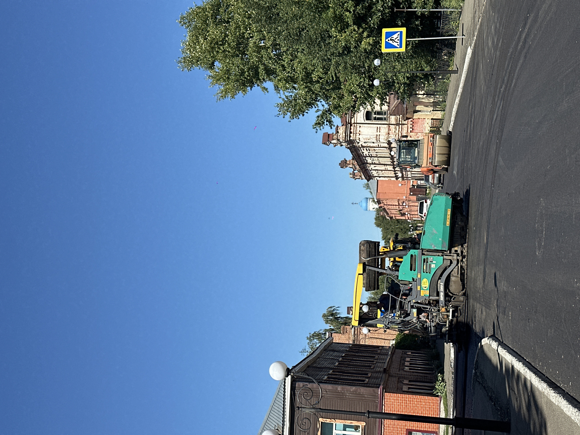 The image captures a street scene in Biysk, Russia, where road construction is underway. A paver, a large green and yellow machine, is actively laying asphalt on the road surface. In the background, a compactor, an orange roller, is visible, likely working to smooth the newly laid asphalt. Several buildings line the street, showcasing varying architectural styles. To the left, a two-story wooden building with red brick detailing stands prominently. Further down the street, an ornate brick building with intricate architectural features is present. A pedestrian crossing sign is posted on the right side of the road, indicating a crosswalk. The sky is clear and blue, suggesting a sunny day. The overall environment is that of an active urban construction site.