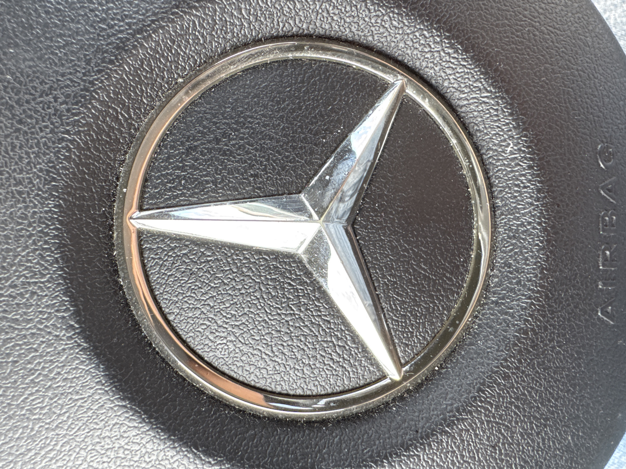 The image is a close-up, eye-level shot of a Mercedes-Benz logo embossed on a black, textured steering wheel, likely in a vehicle. The logo, a silver three-pointed star within a silver circle, is centrally located and prominently featured. The textured black surface of the steering wheel surrounds the logo, with subtle lighting and shadows highlighting its contours. Below the logo, the word "AIRBAG" is faintly visible in uppercase letters, also embossed on the steering wheel. The image appears to be taken indoors or in a shaded area, given the quality of light. The specific location of the vehicle in Folsom, United States is not discernible from the image itself.