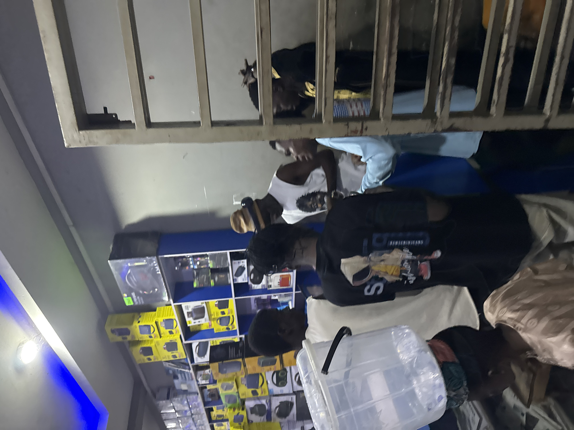 The image depicts a group of people in what appears to be a store or workshop setting, possibly in Jalingo, Nigeria. The scene is dimly lit, suggesting it could be evening or indoors with artificial lighting. The subjects are positioned in a way that makes it difficult to discern a specific activity, but they seem to be gathered around or looking at something.
One individual is wearing a black t-shirt with some visible text and graphics, including the letter "S" and what looks like a basketball player graphic. Another person is wearing a light blue shirt. There are shelves filled with various boxed items, predominantly in yellow and blue packaging, suggesting electronics or similar merchandise. A clear plastic container is also visible in the foreground.
The overall impression is one of casual interaction within a commercial or storage environment. The orientation of the image is skewed, making it challenging to fully interpret the spatial relationships and actions of the individuals. There are no clear indications of time of day or weather conditions, beyond the low light suggesting it's not daytime outdoors. The emotions of the individuals are not clearly discernible due to the image quality and lighting.