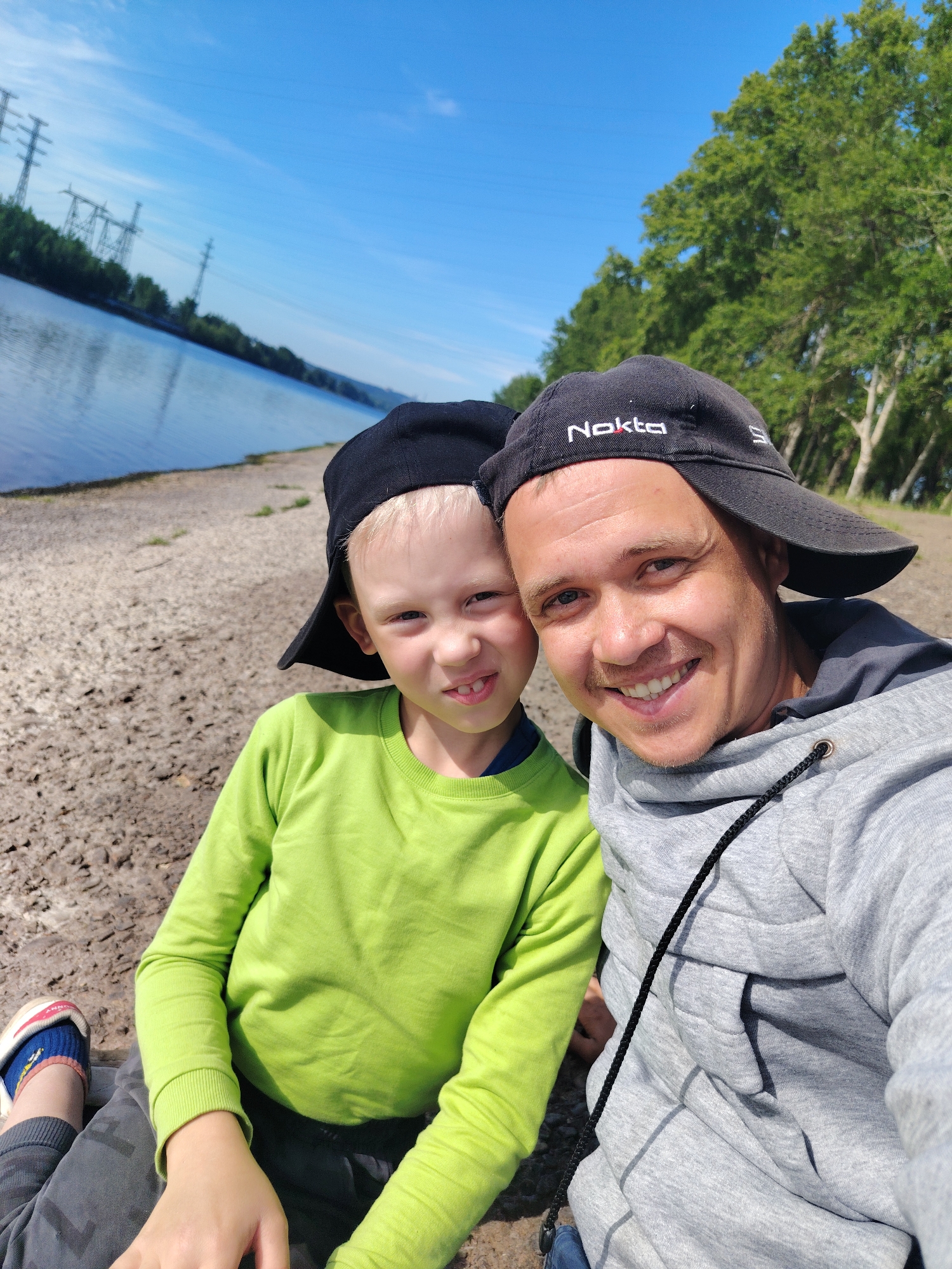 A bright outdoor selfie captures an adult man and a young boy, likely father and son, smiling warmly at the camera. Both subjects wear backward baseball caps; the man's black cap prominently displays "Nokta" in white and red text. The man is dressed in a grey hooded sweatshirt with a black drawstring, while the boy wears a vibrant lime-green long-sleeved shirt and dark trousers, with a blue and white shoe visible.
They are positioned on a sandy or gravelly shoreline beside a wide body of water, which appears to be a river or large lake, reflecting the clear blue sky. The opposite bank is lined with lush green trees. In the background, several large power line towers are visible in the distance, indicating developed infrastructure. The sky is a brilliant blue with a few scattered white clouds, suggesting a sunny day, likely during late morning or afternoon. The smiling faces convey a sense of happiness and enjoyment of their time together outdoors in Chaikovsky, Russia.