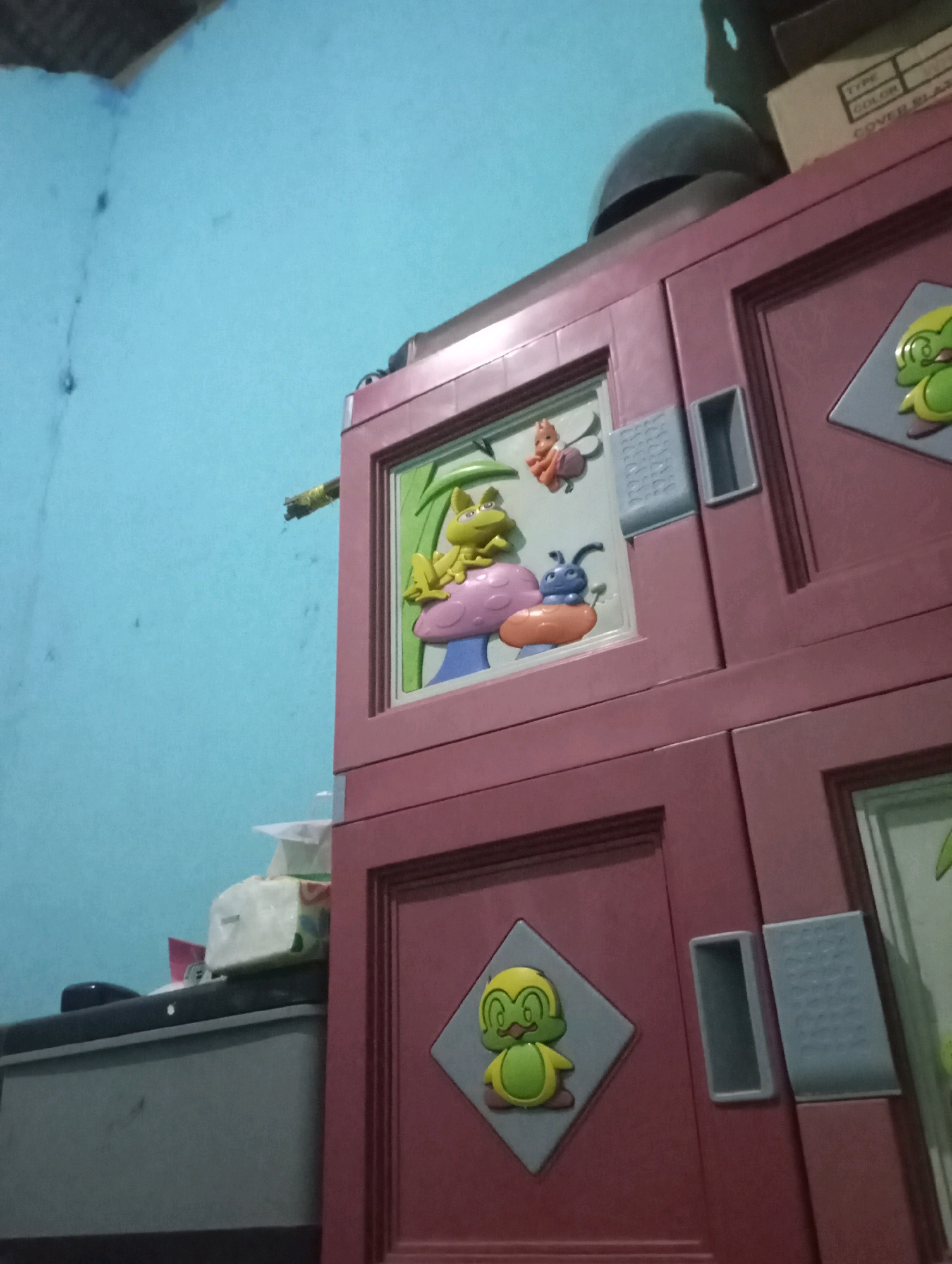The image shows a close-up of a purple plastic cabinet with colorful cartoon characters on its doors. The cabinet is decorated with a frog, a caterpillar, and a bird, suggesting it is a child's furniture. The background is a light blue wall, possibly in a room. There is also a glimpse of what appears to be a gray cabinet or dresser on the left side of the image. The overall impression is of a domestic setting, likely a child's bedroom or play area.
Given the location of Kecamatan Haurgeulis, Indonesia, this scene could be found in a residential home in that area. The specific details visible in the image do not provide any further clues about the exact location within the subdistrict.