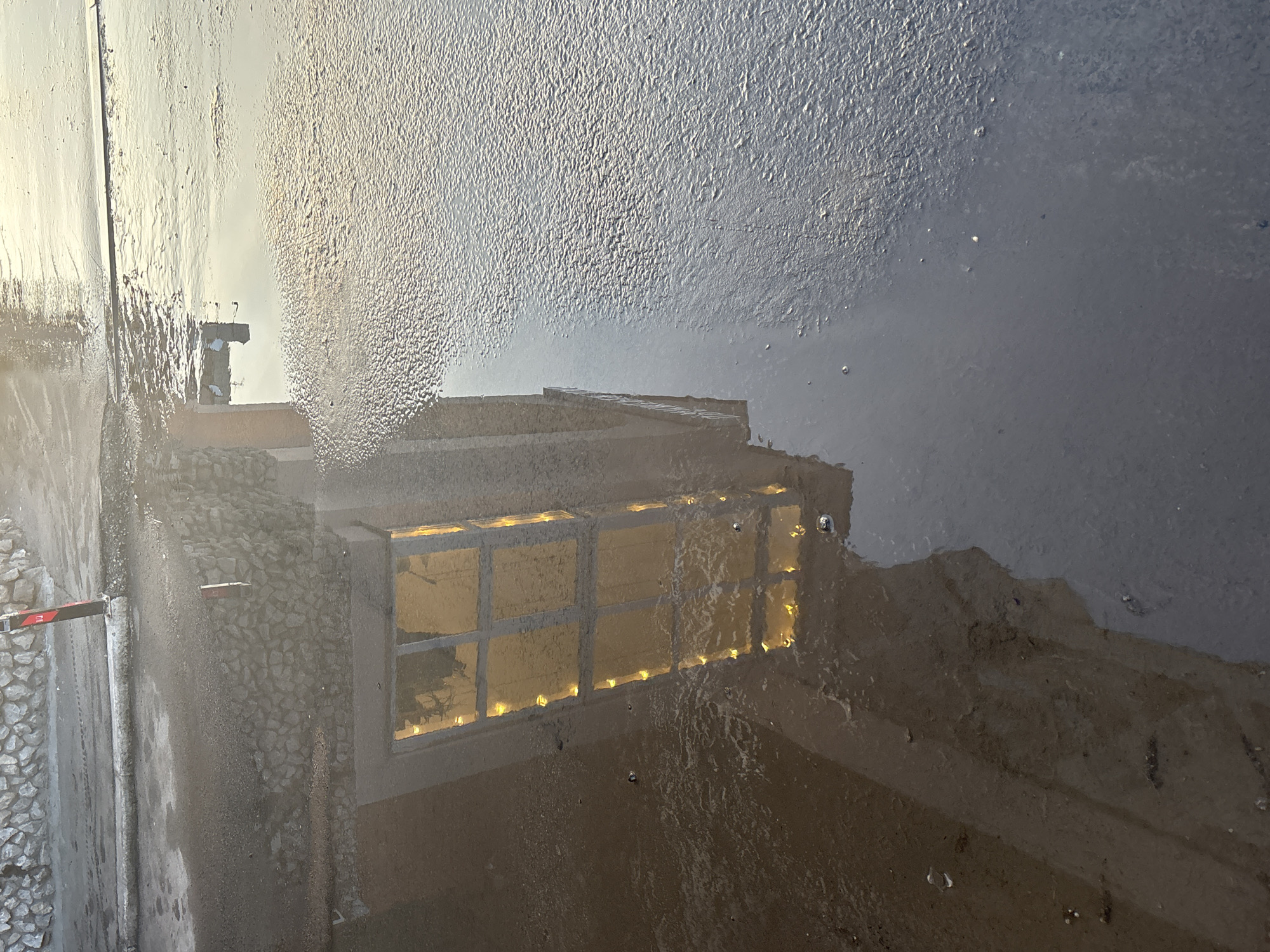 The image appears to show a reflection of a building in a puddle. The building has a prominent window or series of windows illuminated from within. The facade of the building is made of stone or pebbles. The surrounding environment is urban. It is difficult to definitively place it in Perm, Russia without more information.