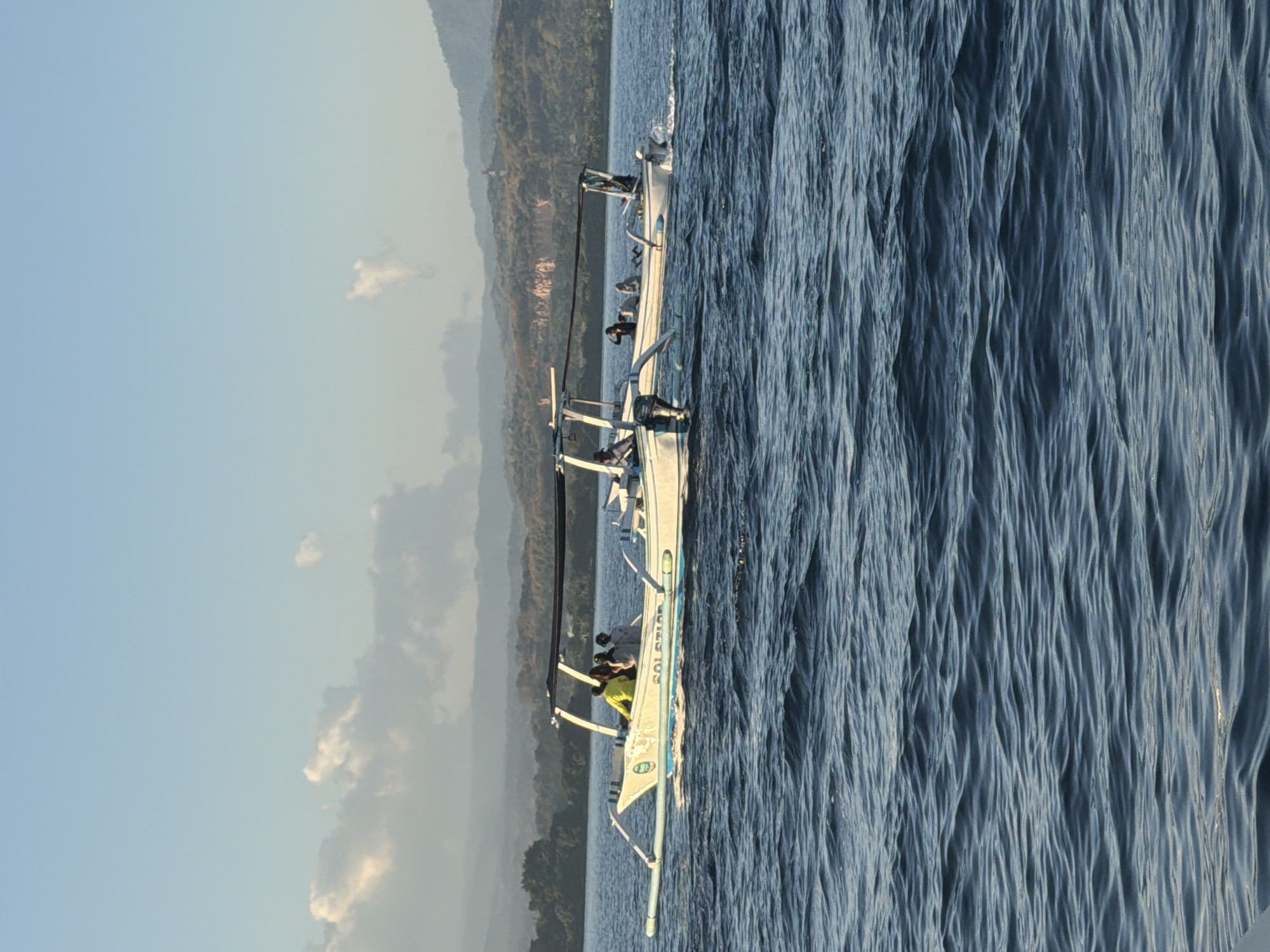 Two traditional outrigger boats are seen on the water in Seririt, Indonesia. The boat in the foreground is white with blue trim and has four people on board. The boat behind it is also white and appears to have three people. Both boats are moving, indicated by the wakes behind them. The water is choppy, and the sky is clear with some scattered clouds. In the background, a lush green coastline with rolling hills can be seen, suggesting a tropical or subtropical environment. The lighting indicates it is either early morning or late afternoon.